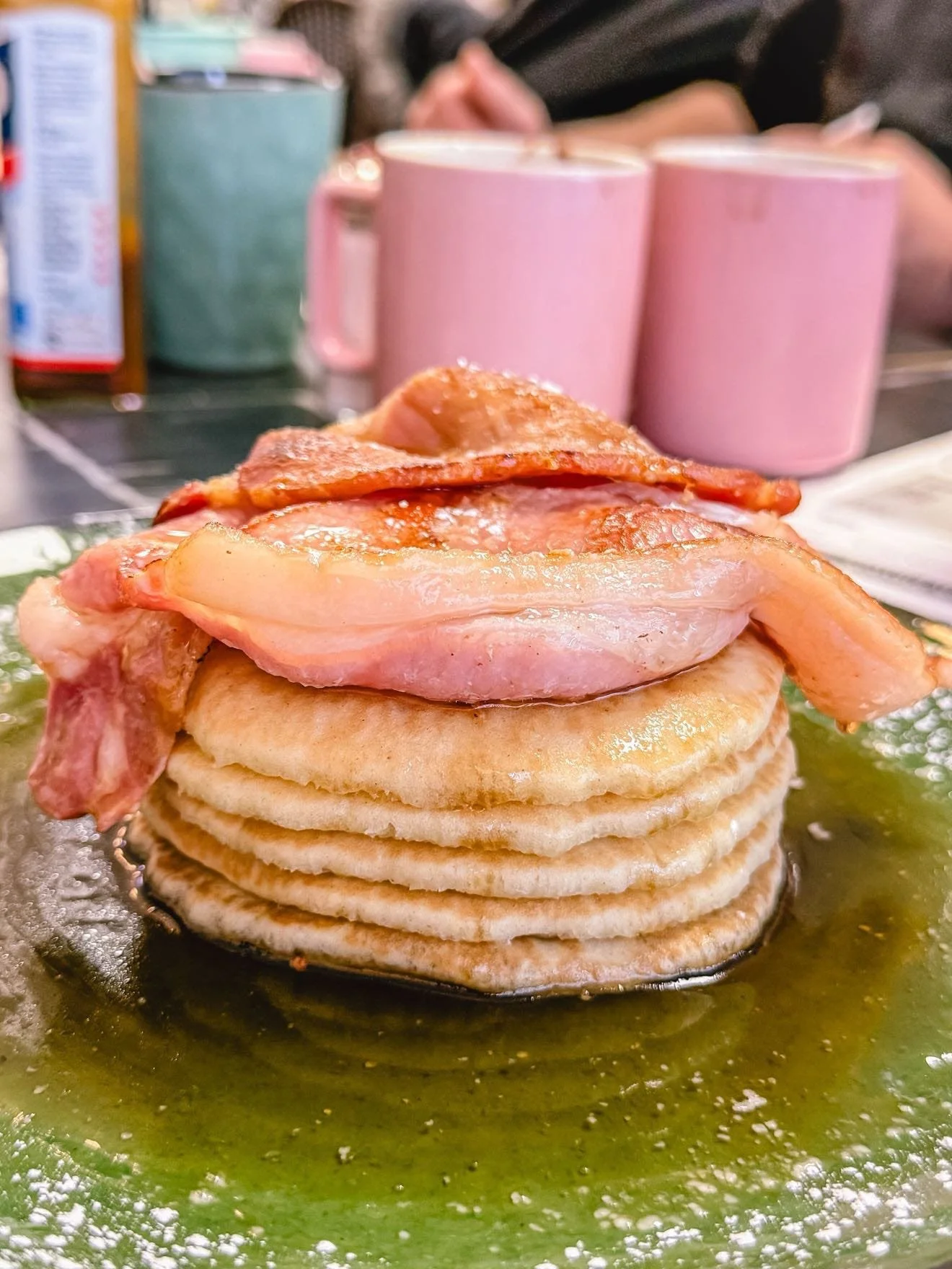 Stack of fluffy pancakes topped with crispy bacon, with syrup around them on a plate.