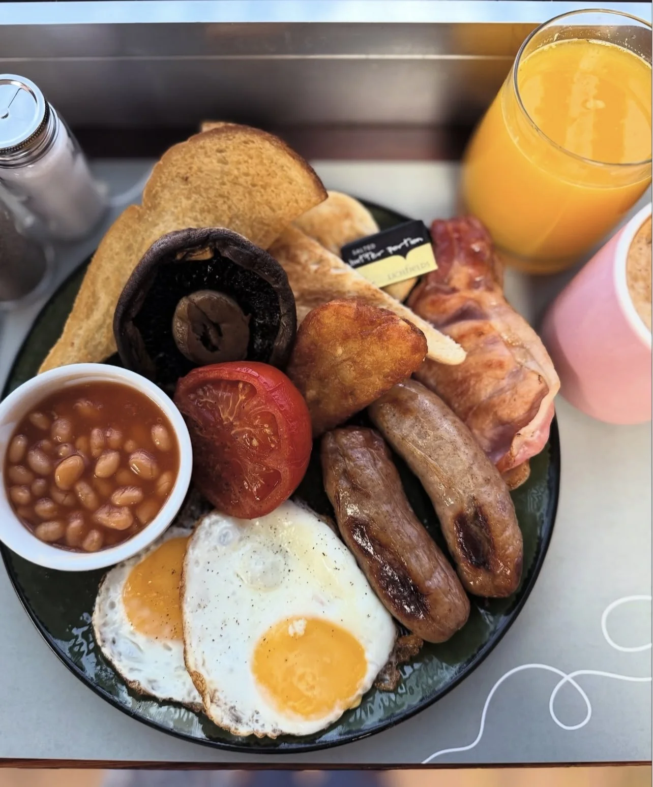 Full English breakfast with fried eggs, sausages, grilled tomato, baked beans, toast, bacon, mushroom, and orange juice.