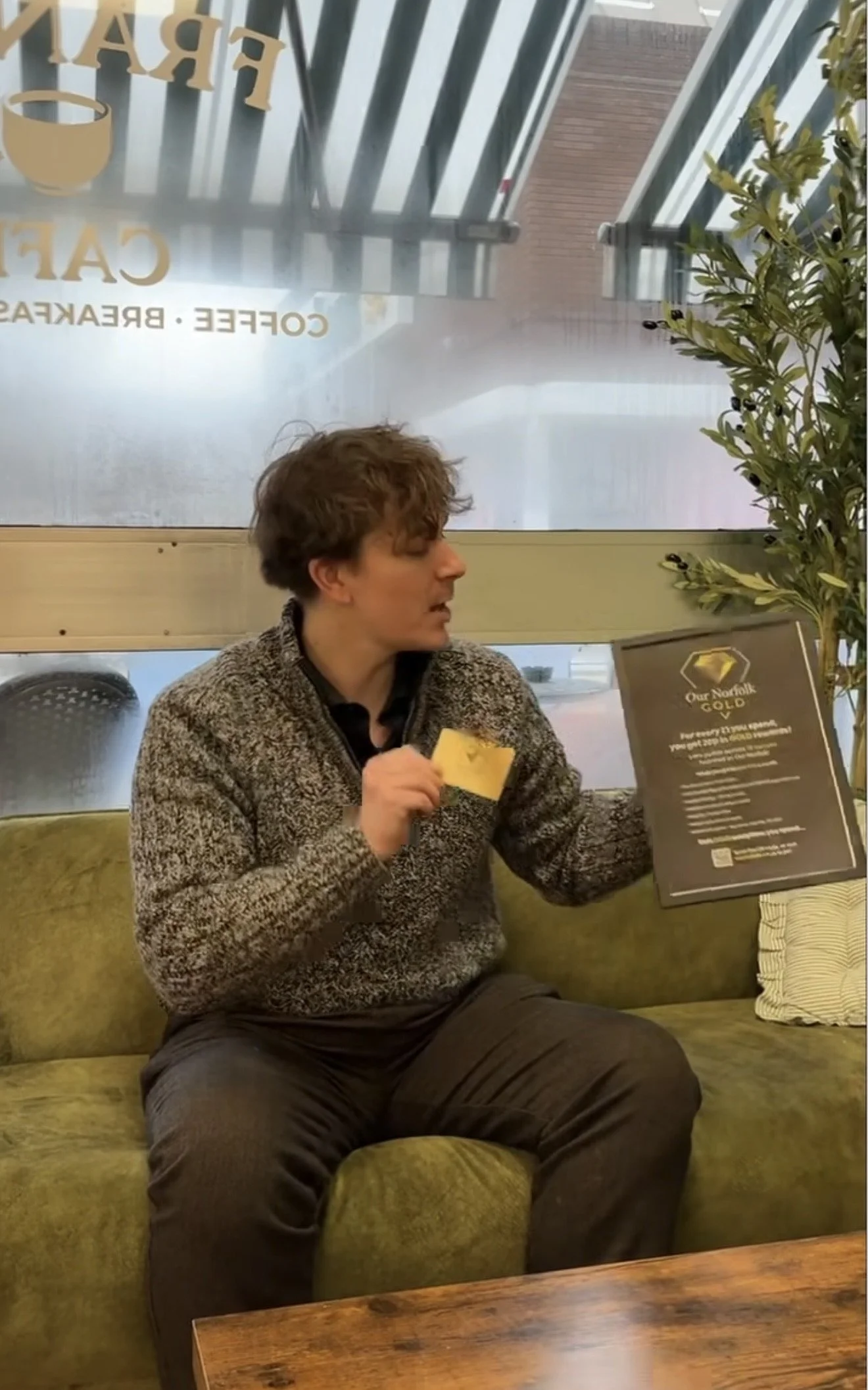 A young man with curly hair sitting on a green sofa in a cafe, holding a yellow piece of paper or napkin in one hand and a menu in the other, with a window behind him showing a street view.