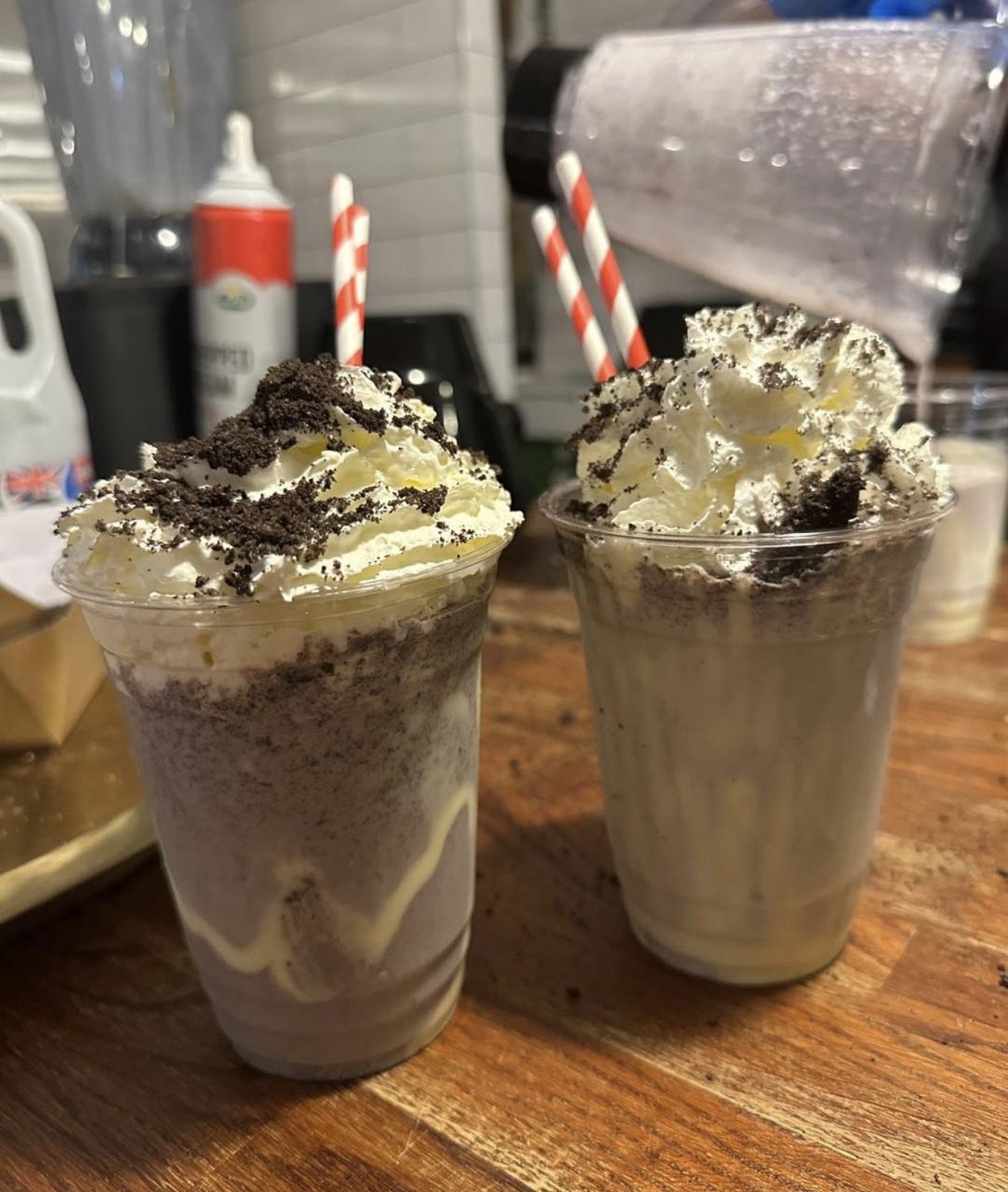 Two cookies and cream milkshakes topped with whipped cream and crumbled cookies, placed on a wooden table with striped straws and background kitchen items