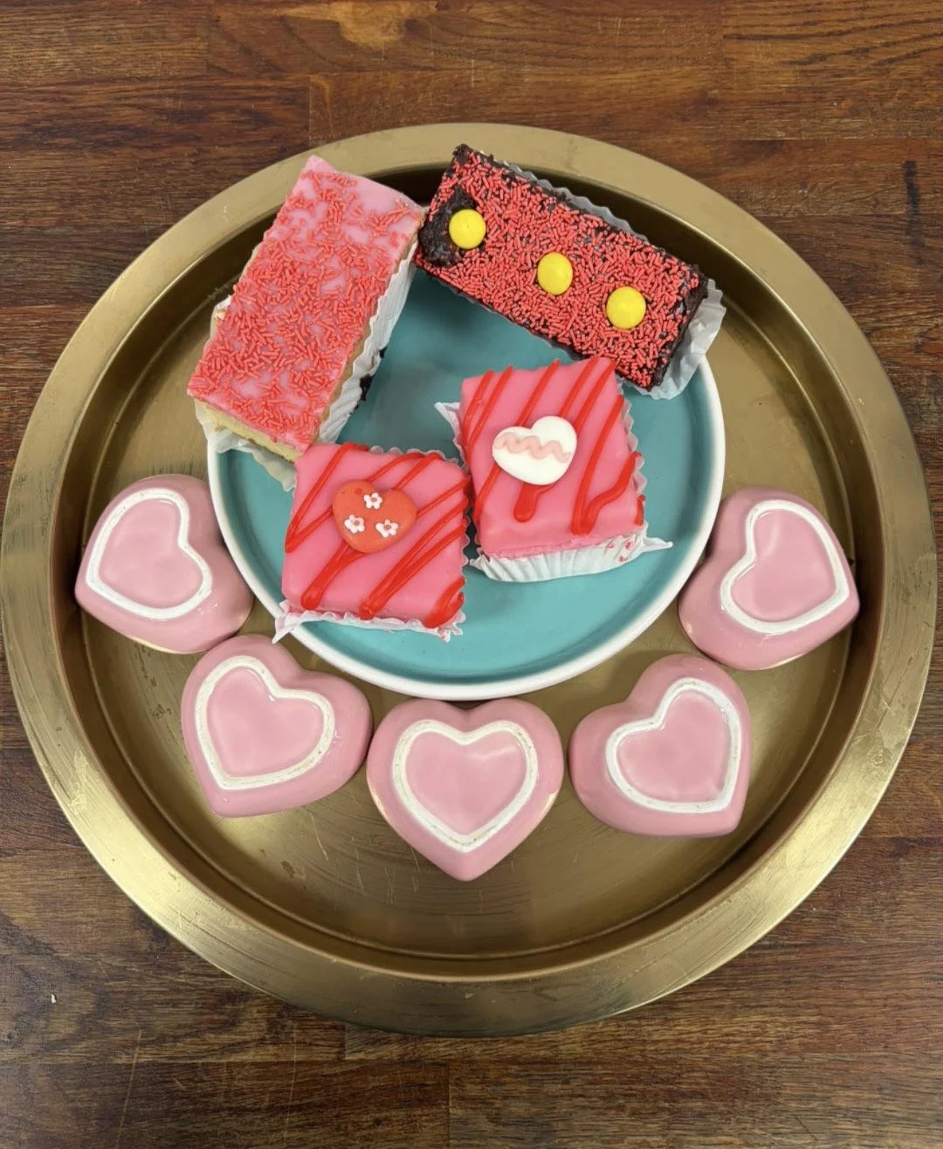 Various decorated Valentine’s Day-themed desserts on a gold tray, including heart-shaped cookies with pink icing and white outlines, and small cakes with pink icing, red drizzle, and Valentine-themed decorations.