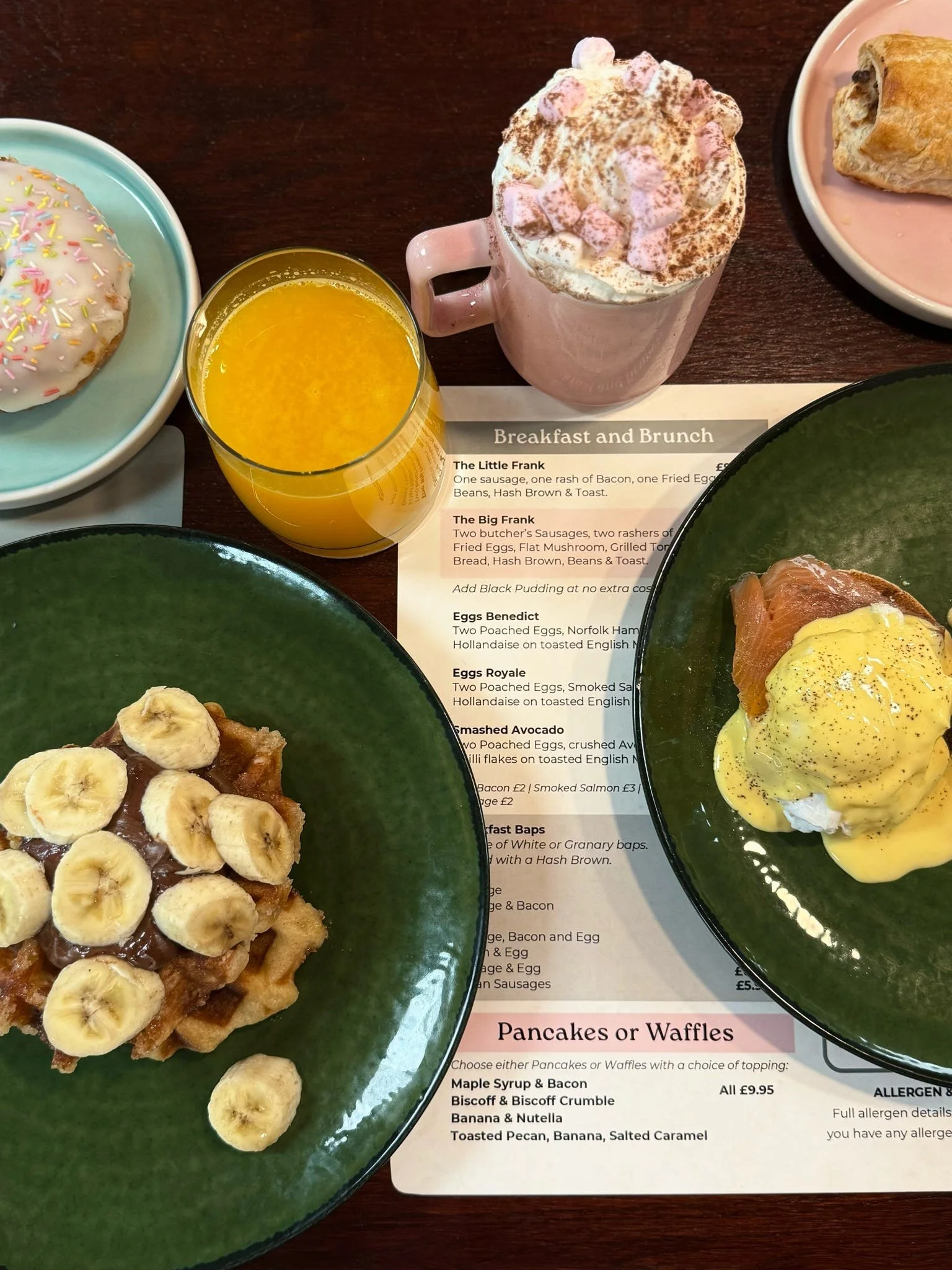 Breakfast with a bowl of banana and chocolate waffles, topped with banana slices and chocolate, a green plate of eggs Benedict with smoked salmon, a green plate with a wrapped croissant, a glass of orange juice, a pink mug of hot chocolate topped with marshmallows, and a pink plate with a croissant.