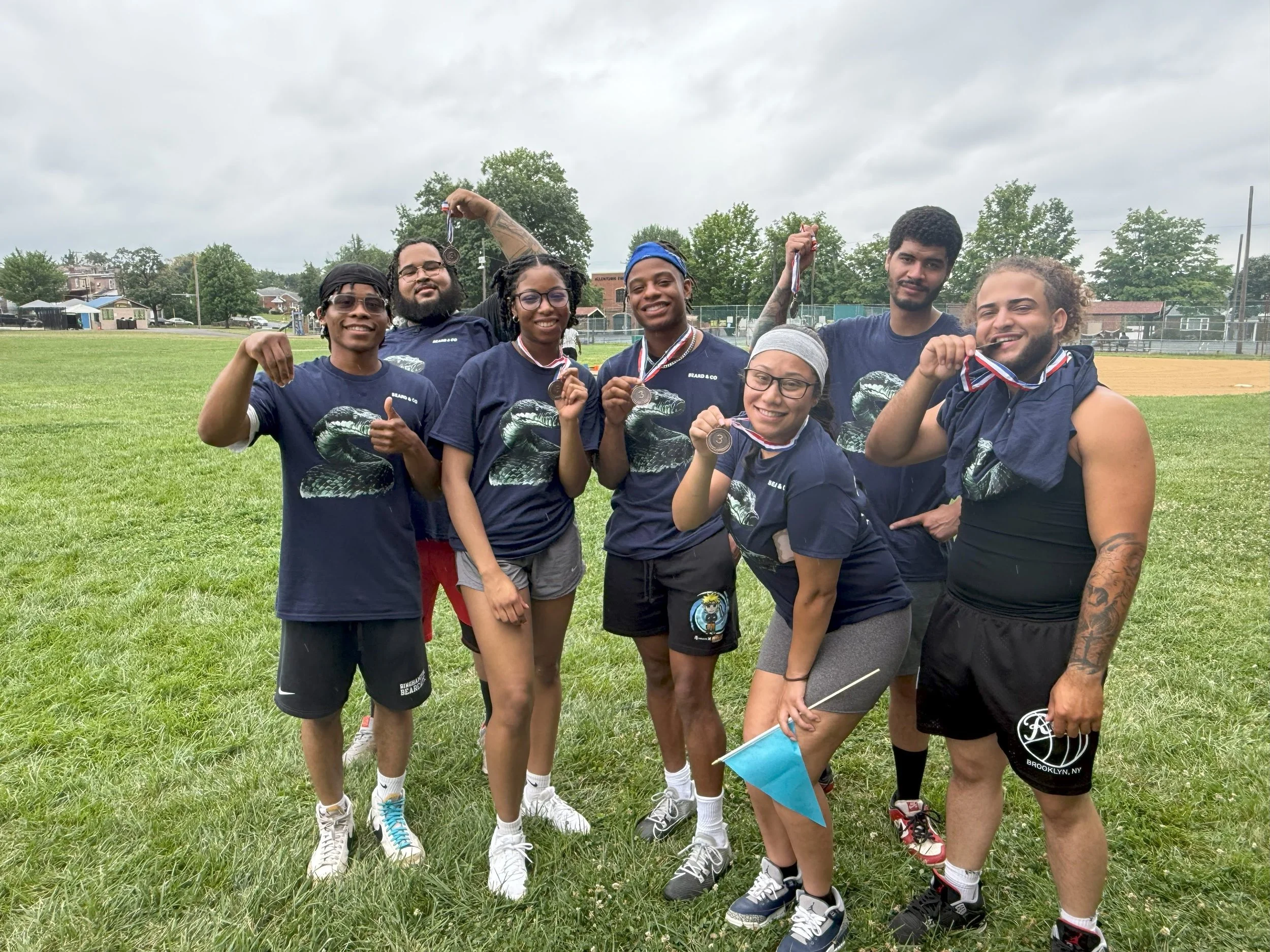 Group of young runners celebrating with medals on a grassy field during an outdoor event on a cloudy day.