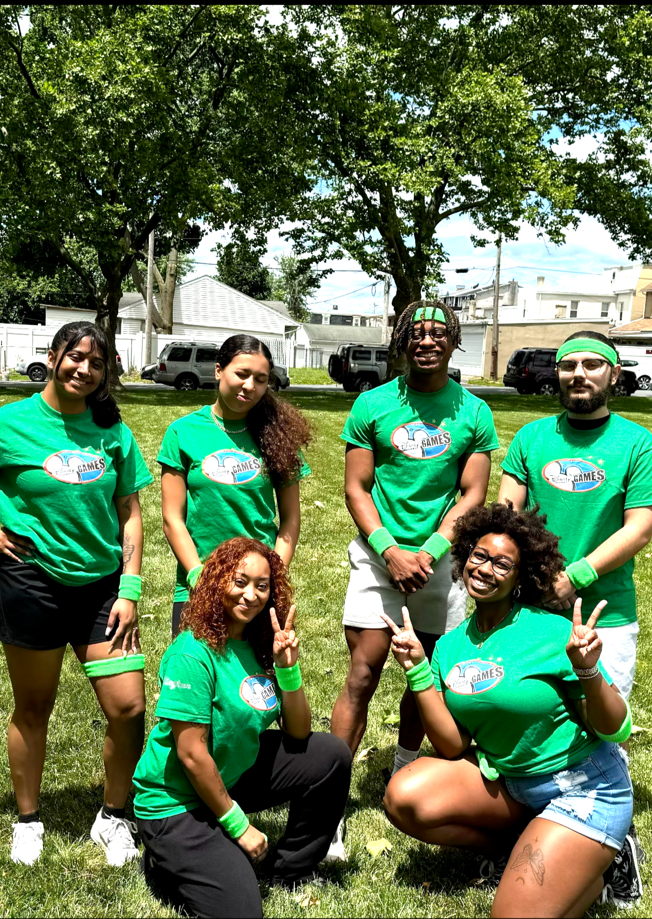 Group of seven people wearing green shirts with a logo, posing outdoors on grass with trees and houses in the background, smiling and making peace signs.