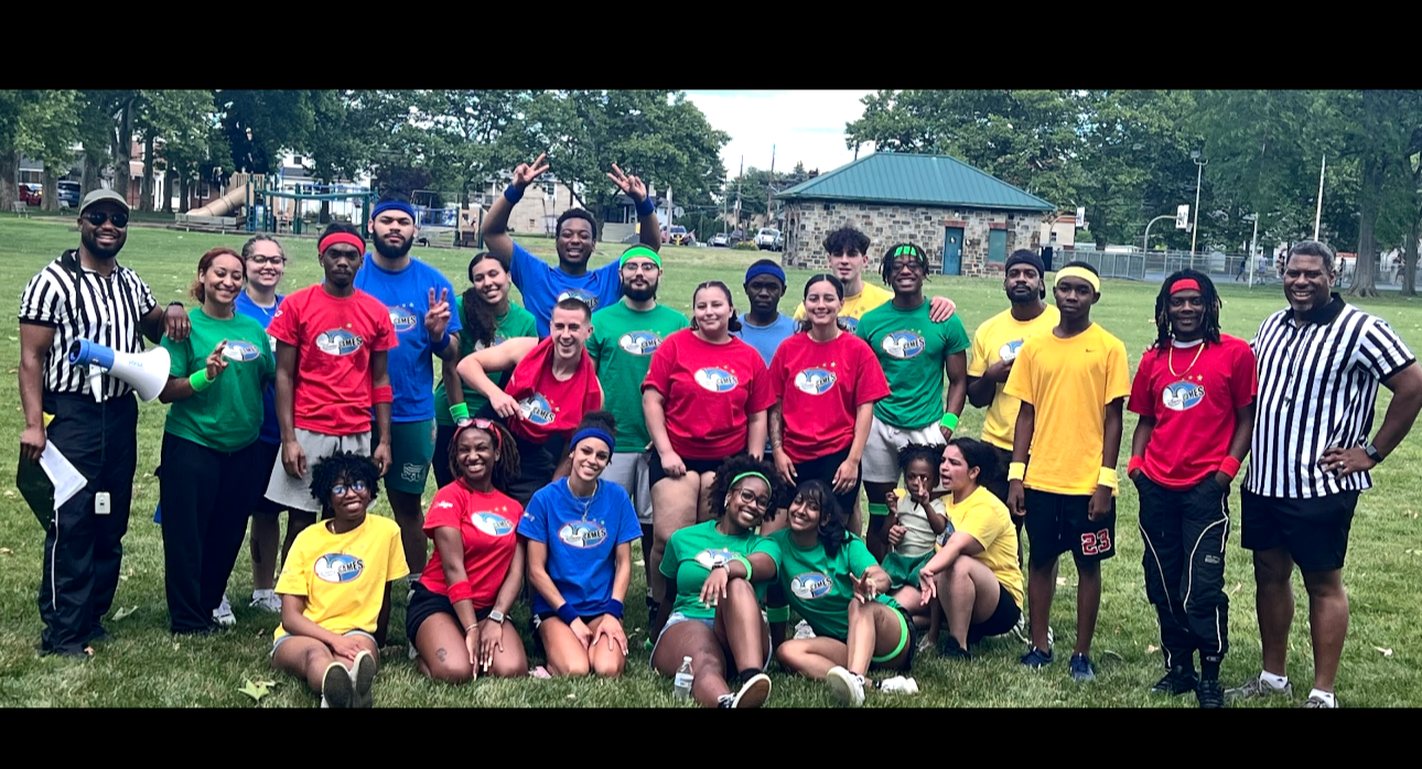 Group of children and adults dressed in colorful athletic shirts, some with yellow, green, red, and blue, posing outdoors on a grassy field with trees and a building in the background. Some are standing, sitting, or kneeling, displaying cheerful expr