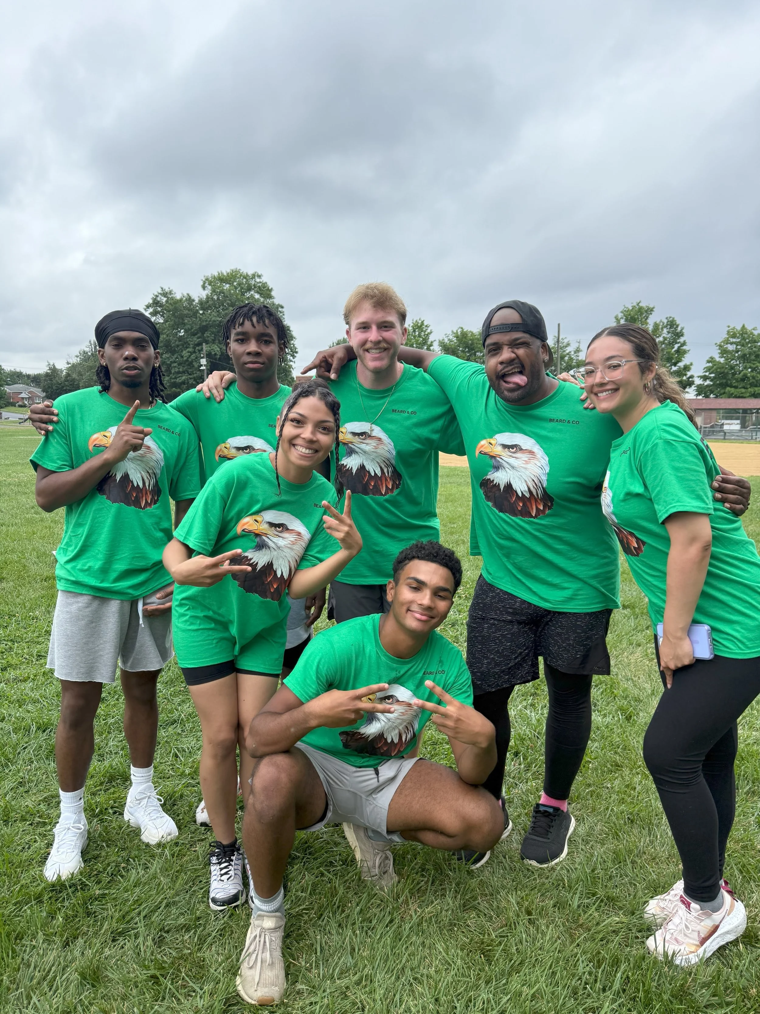 Group of seven young people outdoors on a grassy field, wearing matching green T-shirts with an eagle graphic, smiling and posing for a photo before cloudy skies.