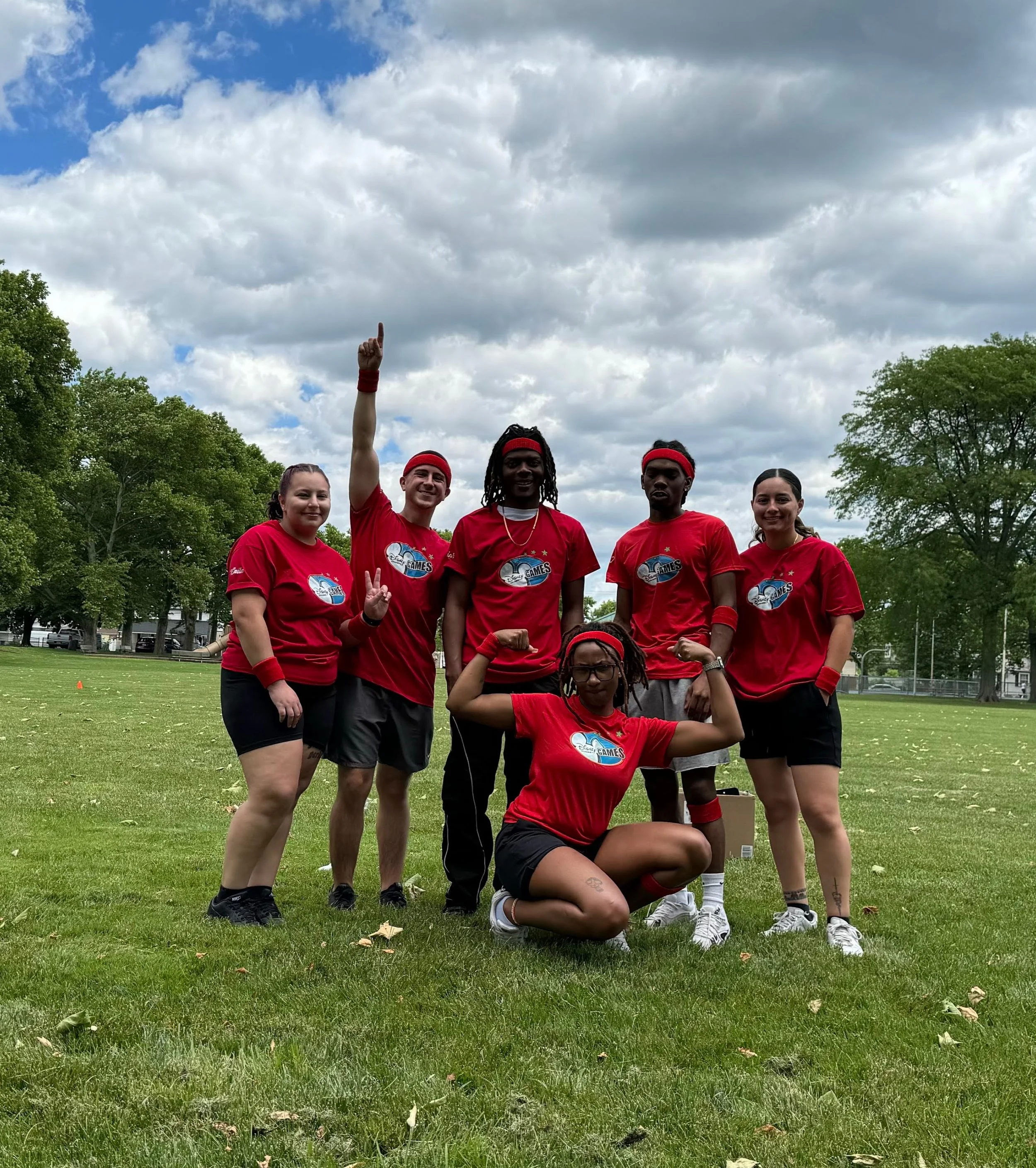 A group of six young people in red athletic shirts posing on a grassy field, with some standing and one kneeling in front, under a partly cloudy sky.