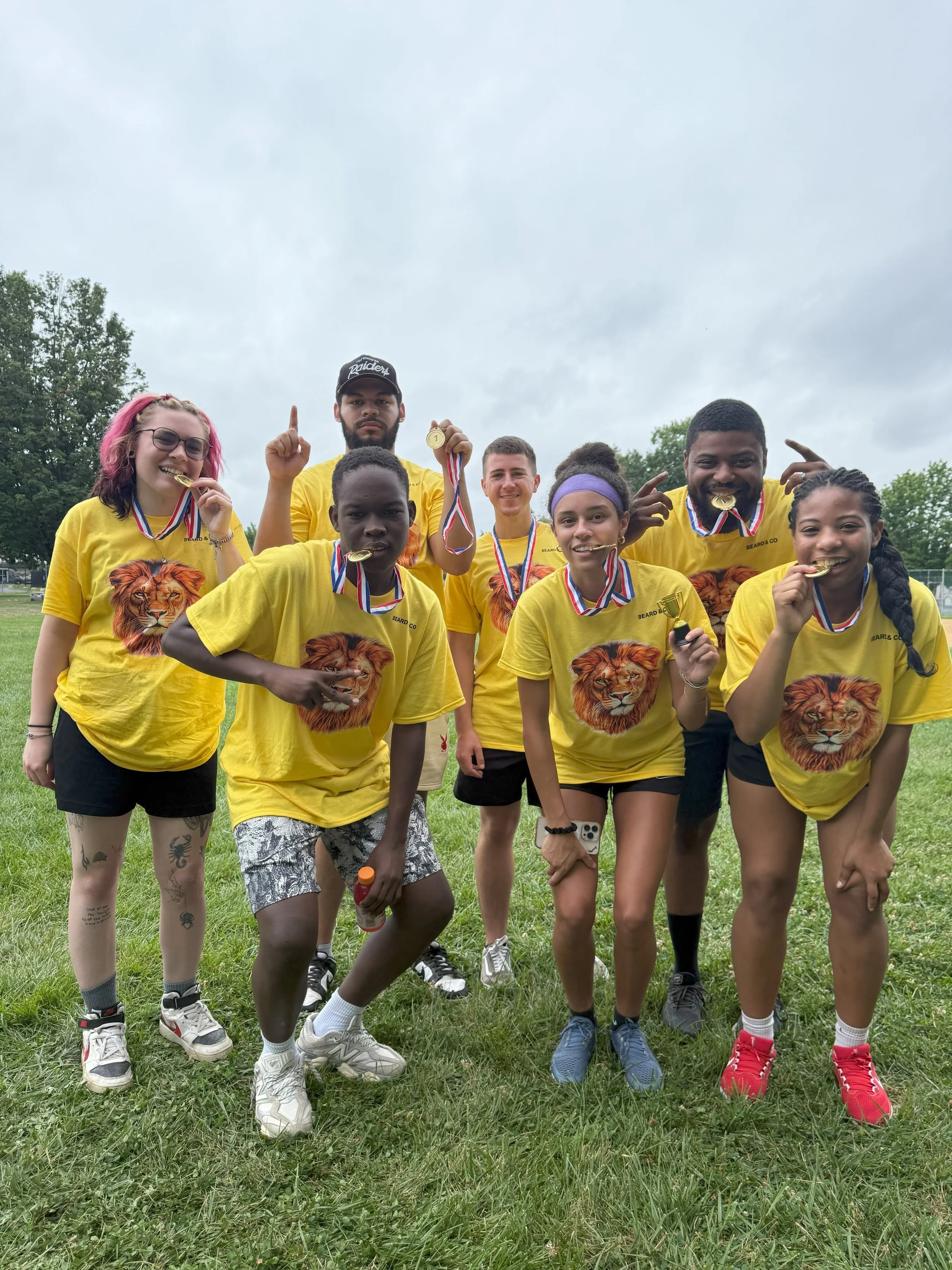 Group of young athletes wearing yellow lion-themed shirts, celebrating and displaying medals in a grassy outdoor area.