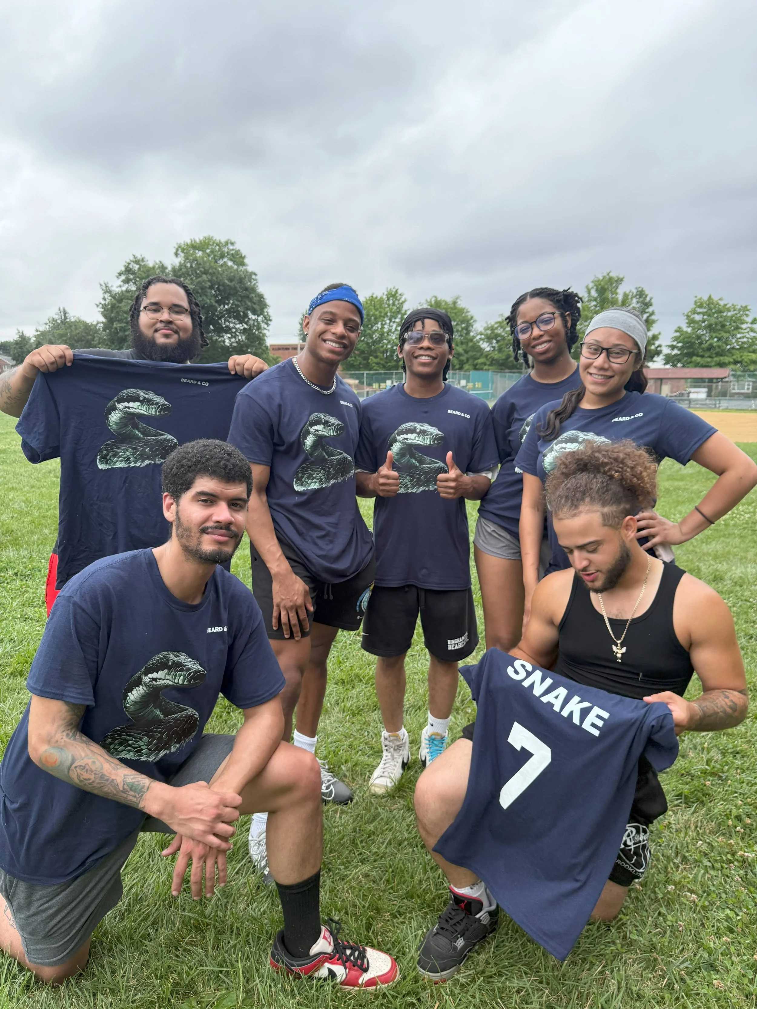 Group of seven young adults, five men and two women, outdoors on a grassy field under cloudy skies, posing with some holding sports jerseys, one with a snake design and another labeled 'SNAKE 7'.