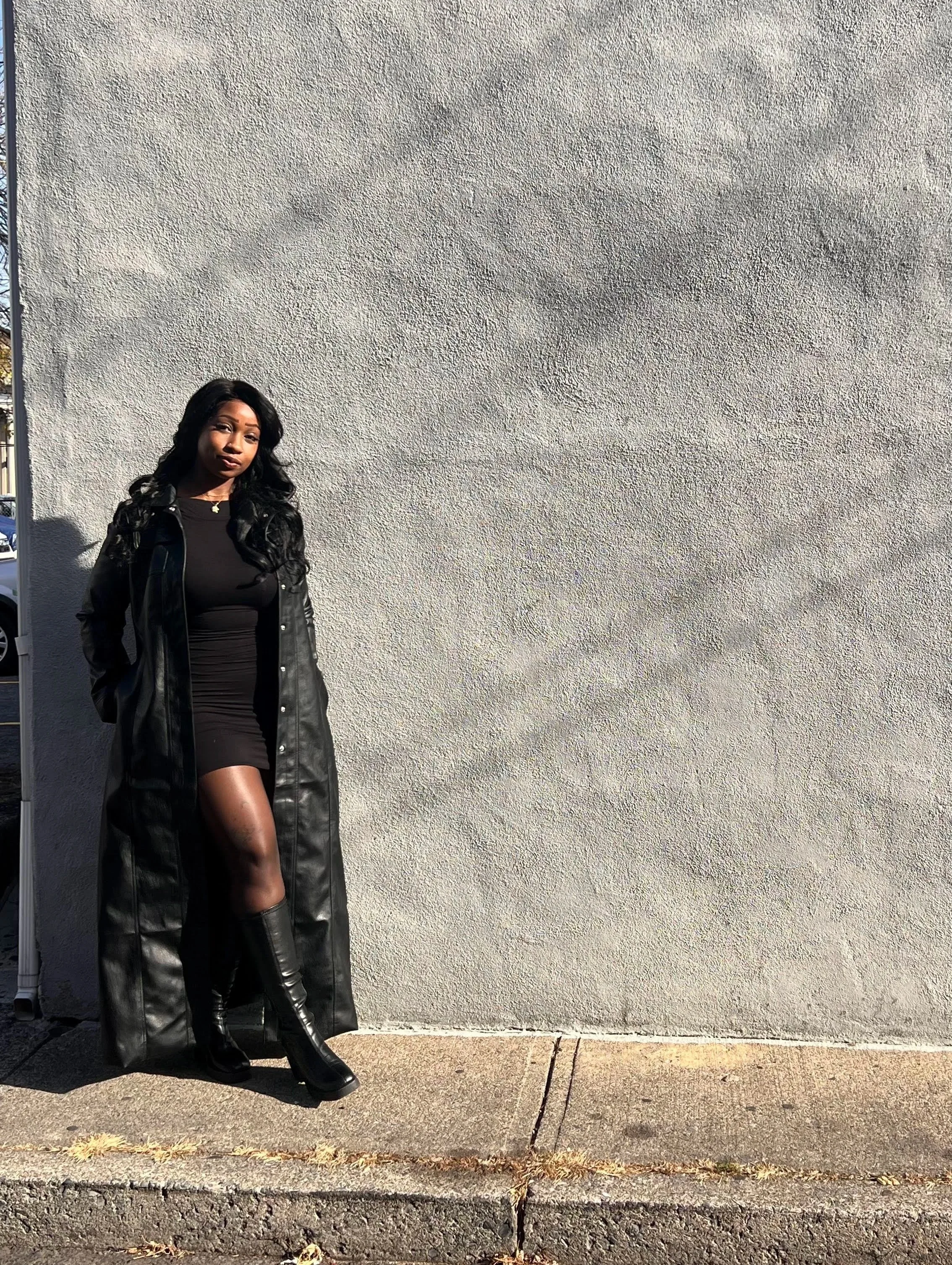 A woman standing against a plain gray wall, wearing a black dress, knee-high black boots, and a long black leather coat, with her hands in her coat pockets.