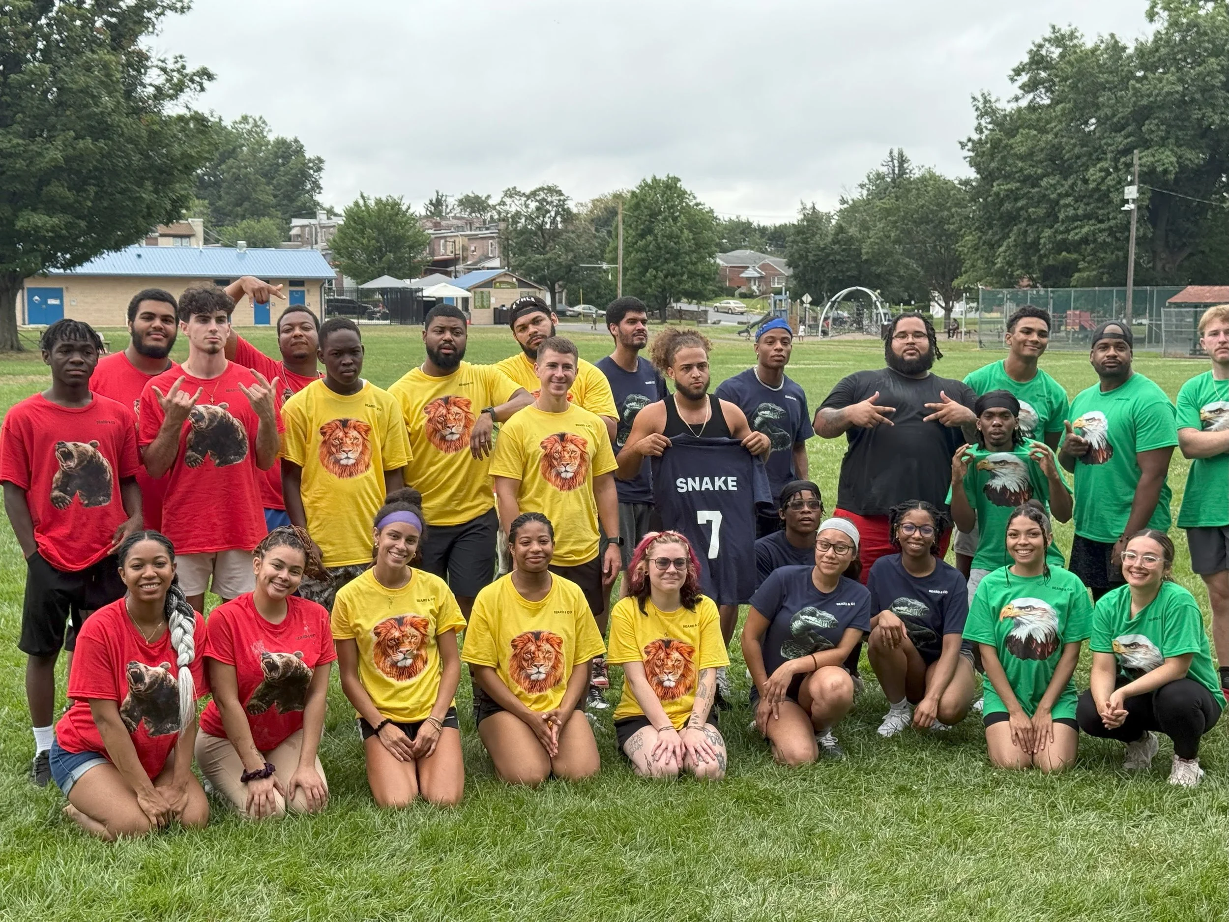 A group of young adults posing outdoors on a grassy field, wearing colorful team shirts with animal prints and a jersey with the number 7. Some are kneeling in front, while others are standing behind them, with a sports playground and trees in the ba