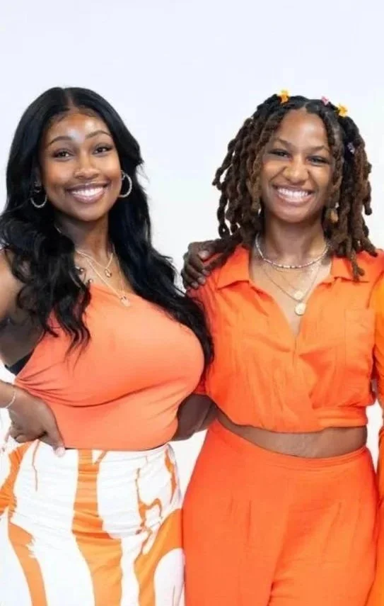 Two smiling women standing close together, one with long black hair and the other with curly hair and colorful hair accessories, both wearing orange outfits, against a plain white background.