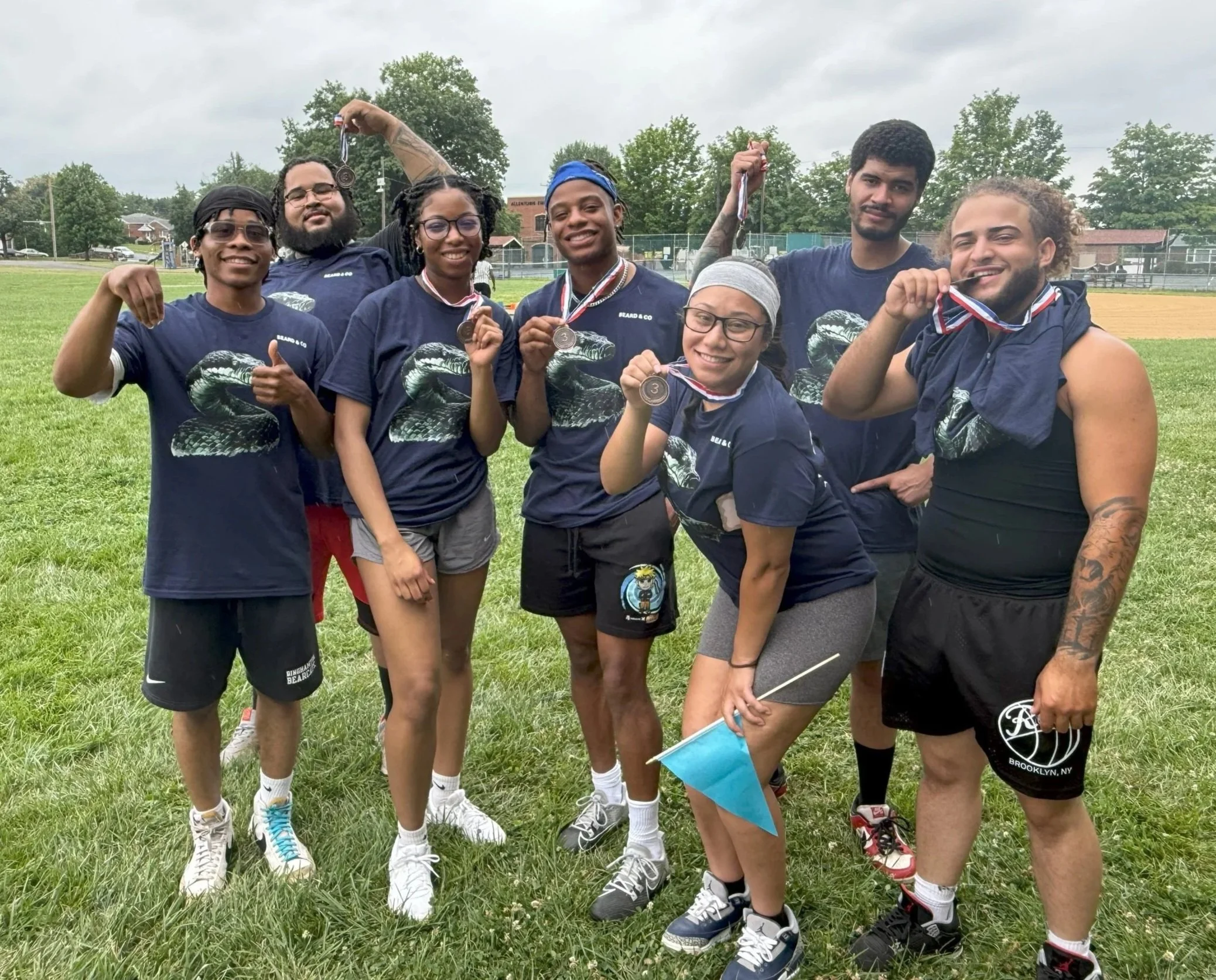 Group of seven young adults outdoors on a grassy field, wearing matching navy blue T-shirts with a snake graphic, holding medals and celebrating a sports event.