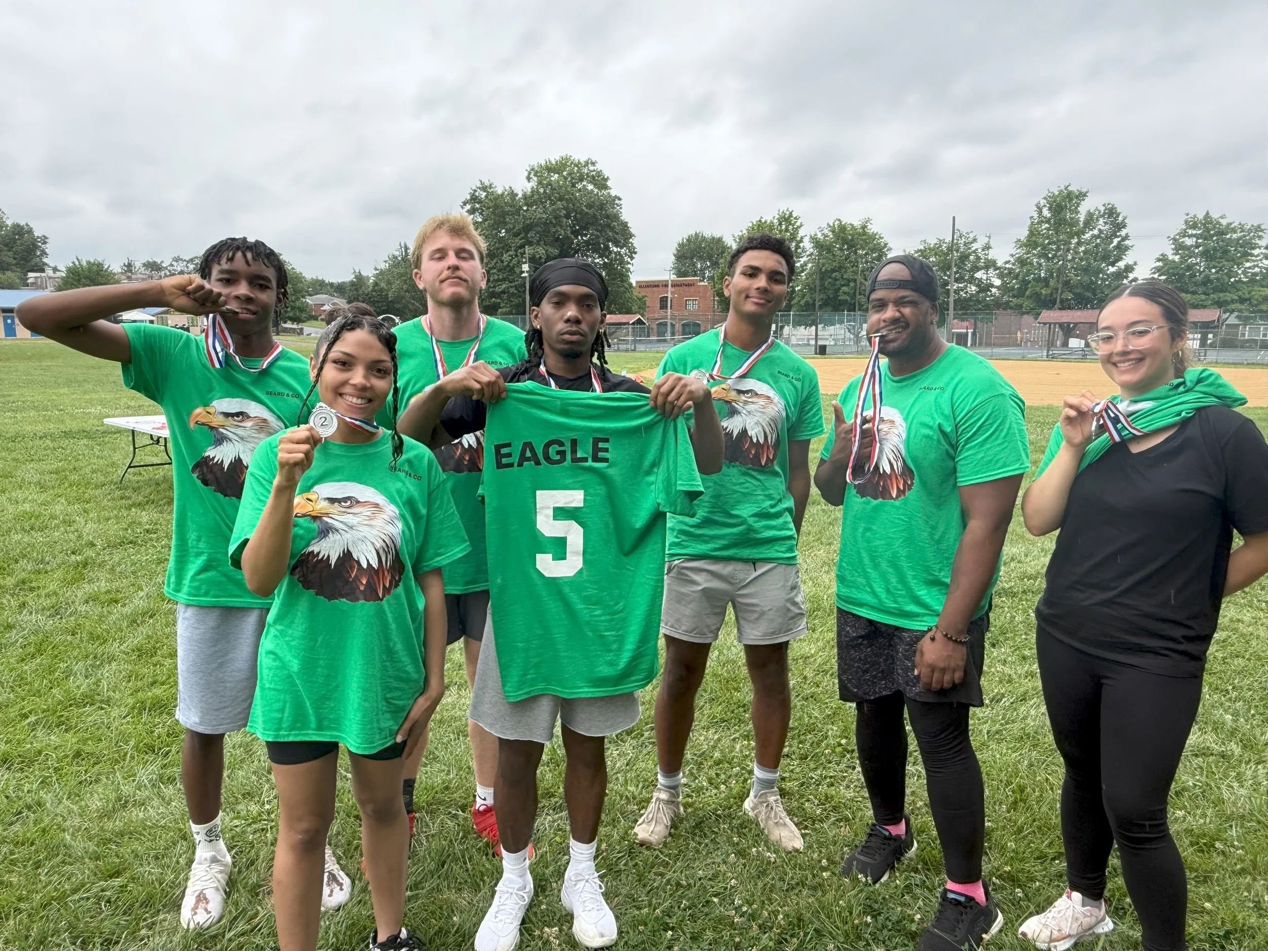 Group of seven young people on a field holding medals and a green sports jersey with number 5, wearing green shirts with an eagle graphic celebrating their sports event victory.