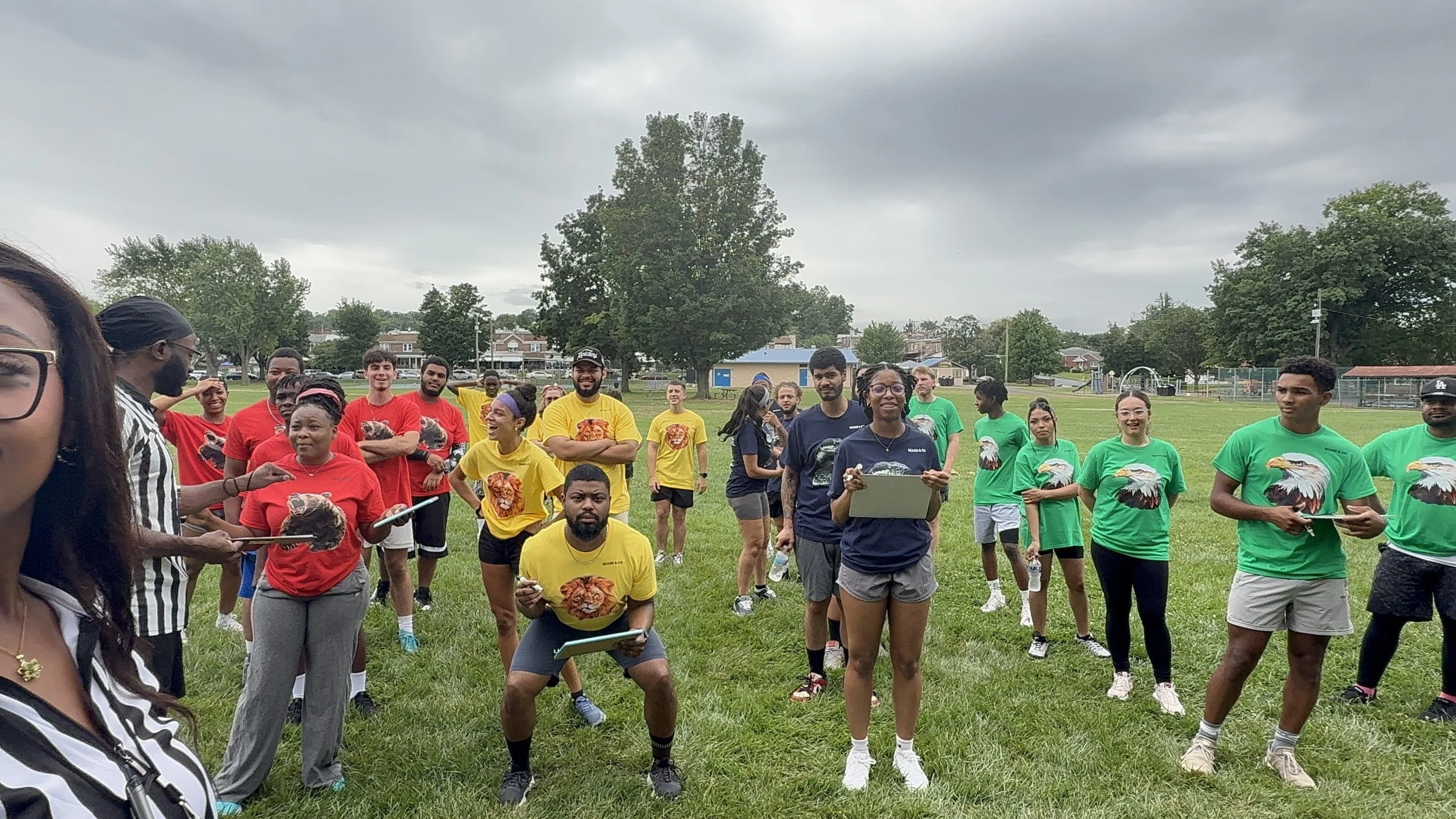Group of children and teenagers gathered on a grassy field for an outdoor activity, some holding clipboards or sheets of paper, dressed in colorful t-shirts with various colors like red, yellow, green, and navy blue, in a cloudy sky setting with tree