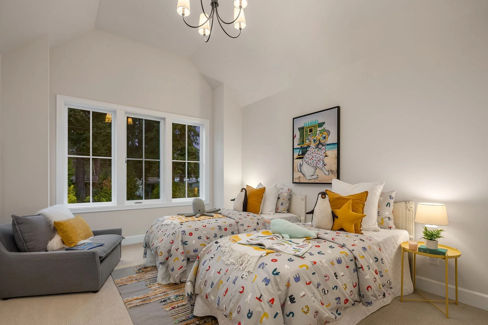A bedroom with two beds covered in white dinosaur-patterned bedding, decorated with plush toys and pillows, large window showing greenery outside, artwork of a dog at the beach hanging above, a gray armchair with pillows and book on the left, a yello