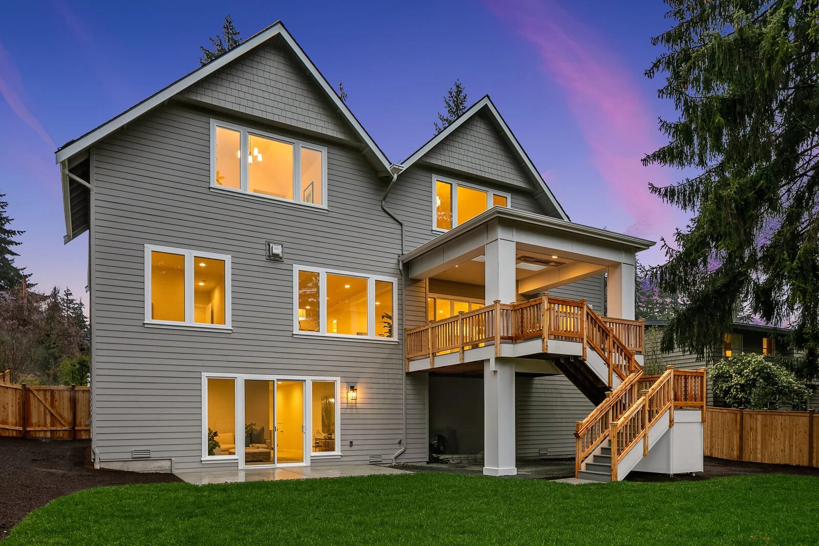 A two-story gray house with white trim and large windows illuminated from within, featuring a wooden staircase leading to an upper porch, set against a evening sky with a purple hue and surrounded by a green lawn and trees.