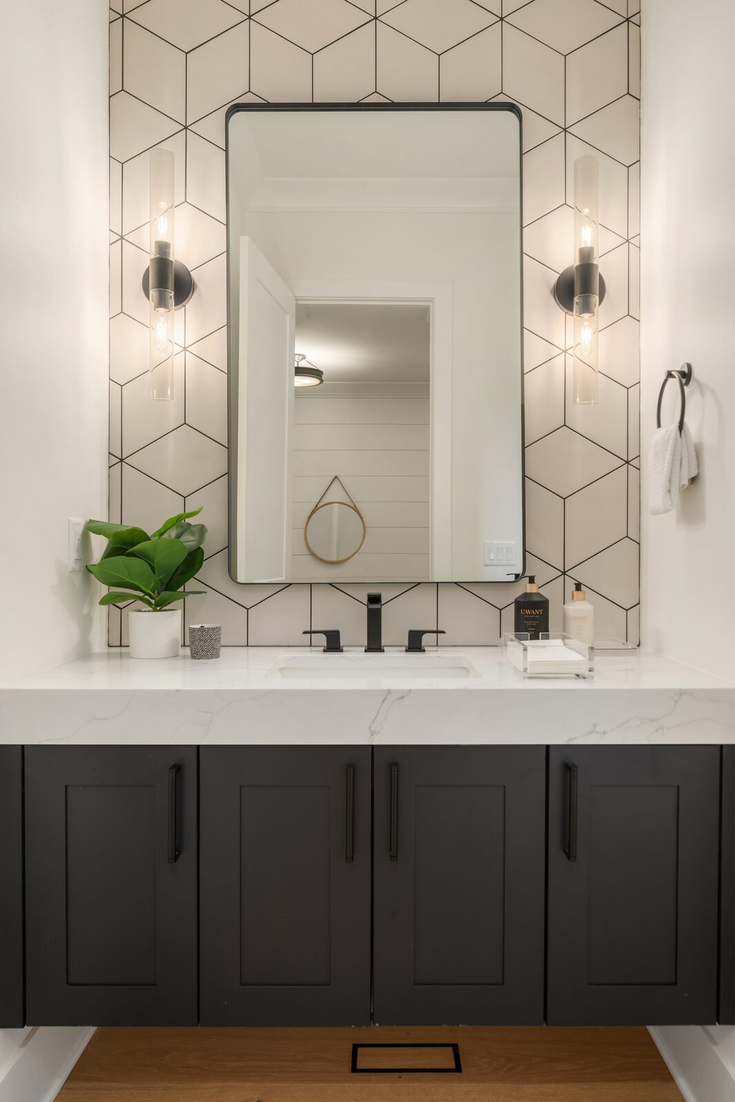 Modern bathroom vanity with a large mirror, black fixtures, decorative black and white wall tiles, a white countertop with a plant on the left, and two wall-mounted light fixtures.