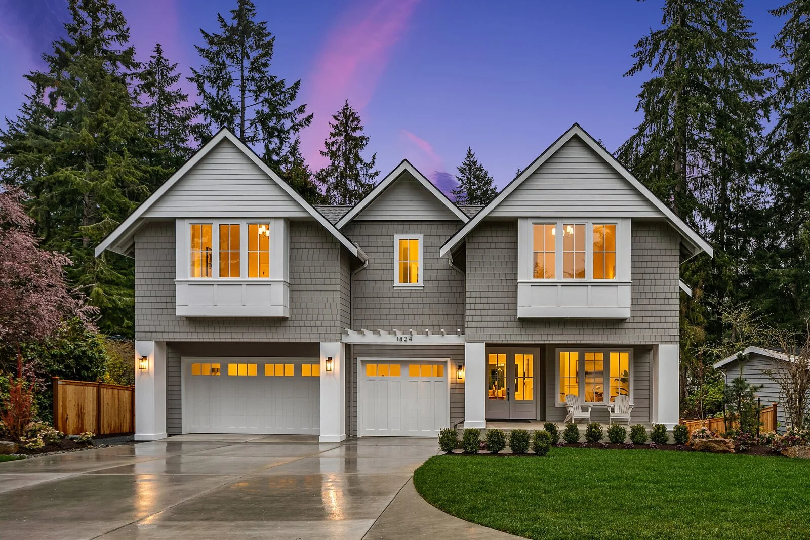 A modern two-story house with gray siding and white accents, illuminated from inside, surrounded by trees and a well-maintained lawn, with a concrete driveway leading to two garages.