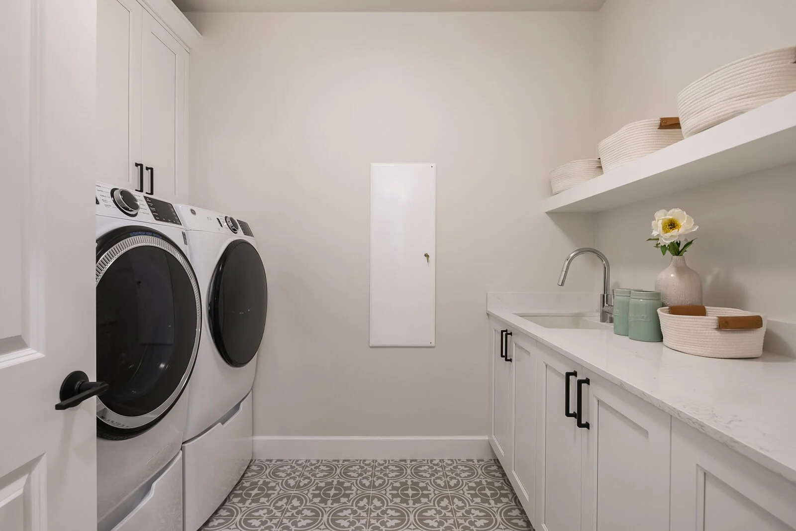 A laundry room with a washer and dryer, white cabinets, a countertop with a sink, decorative bowls, a flower in a vase, and shelves with baskets.
