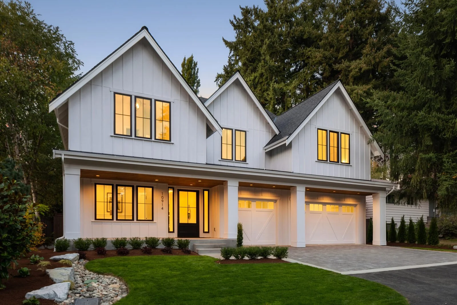 A modern two-story house with white exterior, black-framed windows, and a garage. The house is illuminated from within, with a well-manicured lawn and landscaped garden in the front yard, surrounded by large trees.