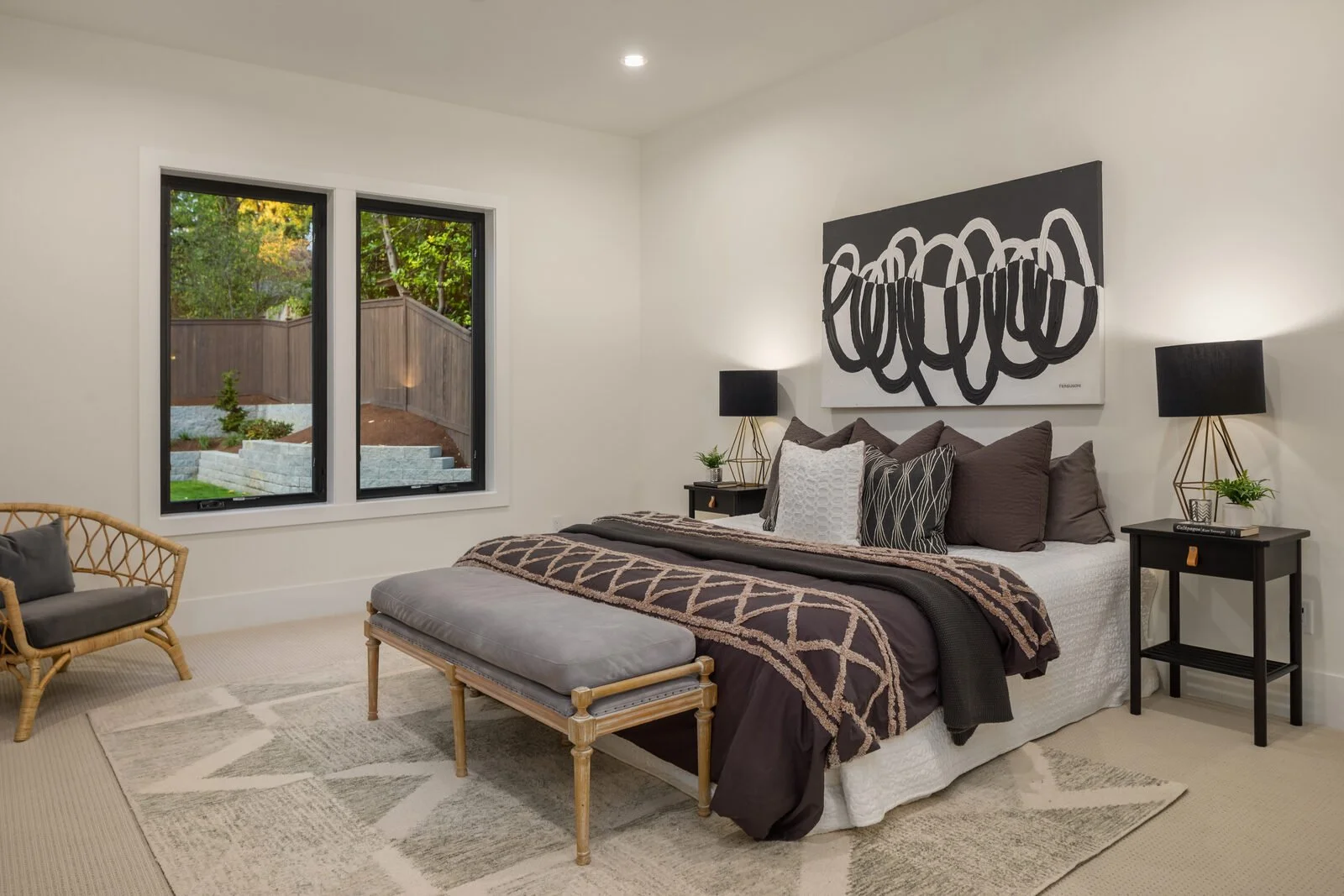 A modern bedroom with a large bed, black and white artwork above, black nightstands with lamps and plants, a window showing an outdoor yard, and a wicker chair with gray cushions.