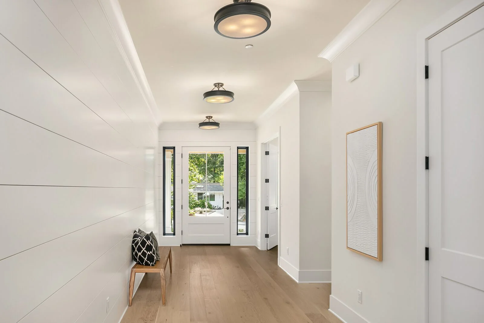 A bright, modern entryway with white walls, a light wood floor, and black ceiling lights. There is a bench with two black and white patterned pillows and a glass door with side windows showing a view of greenery outside.