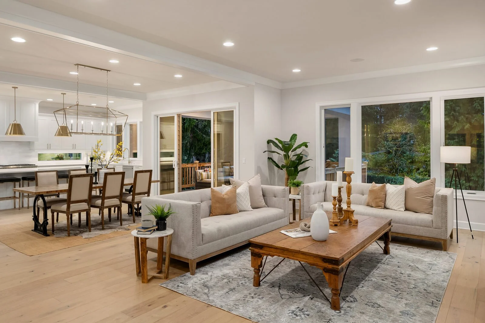 Open-concept living and dining room with white walls, large windows, hardwood floors, and neutral-toned furniture including sofas, a wooden coffee table, and a dining table with chairs, decorated with plants and vases.