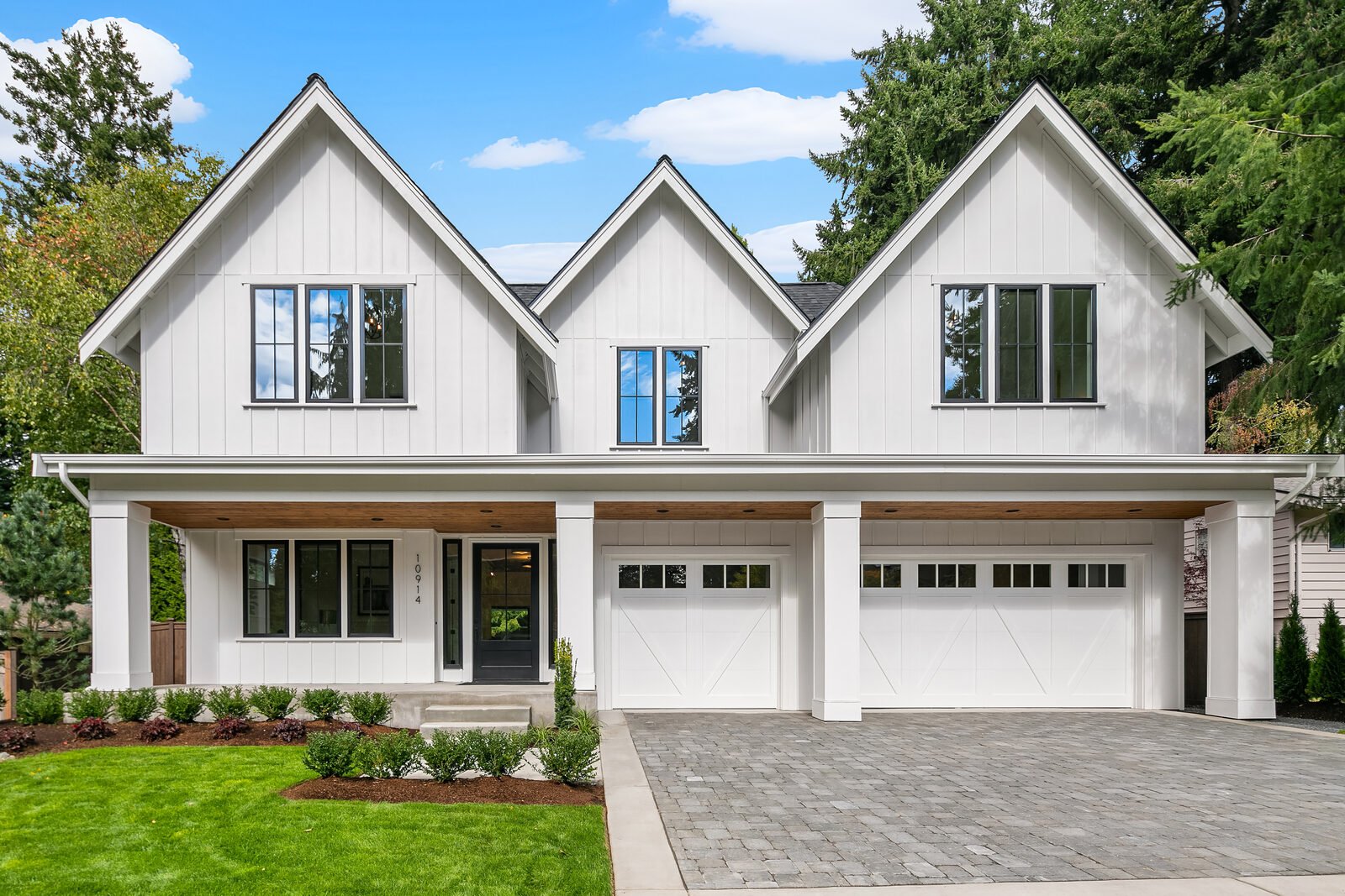 White modern two-story house with black trim, featuring a double garage, front porch, and large windows, surrounded by greenery and a paved driveway.
