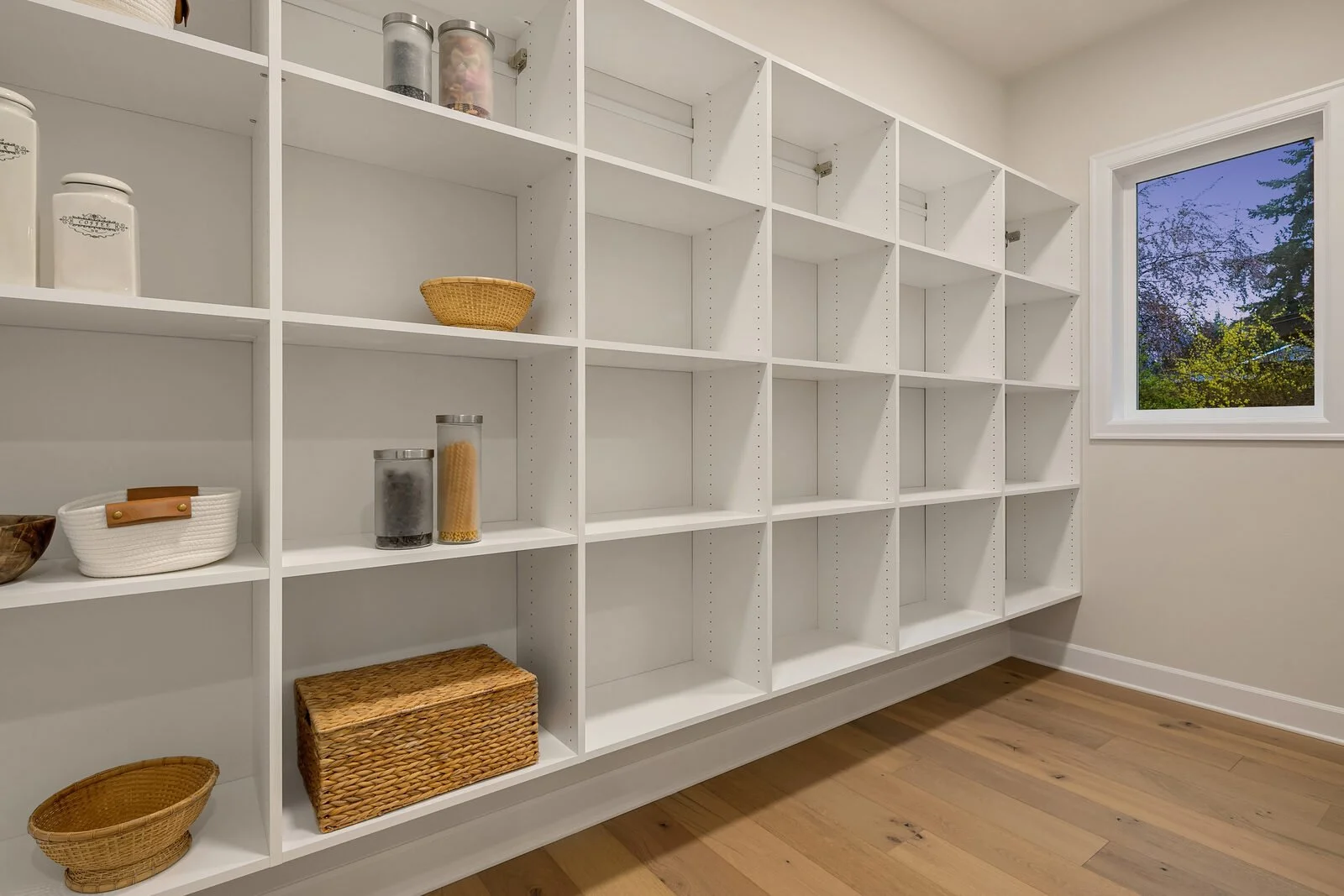 Empty white shelving unit in a room with hardwood floors and a window showing trees outside.
