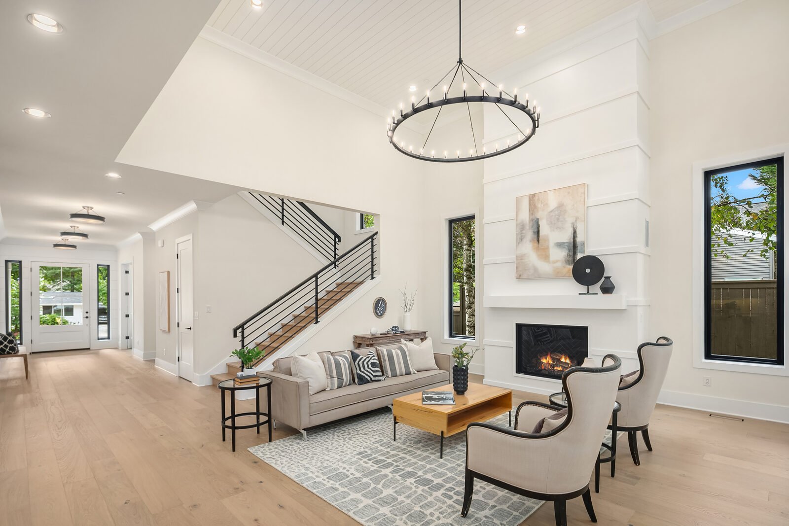 Bright, modern living room with white walls, hardwood floors, a fireplace, and large windows. Features furniture including a beige sofa with pillows, two armchairs, a wooden coffee table, and a small side table, with decorative art on the wall and a 