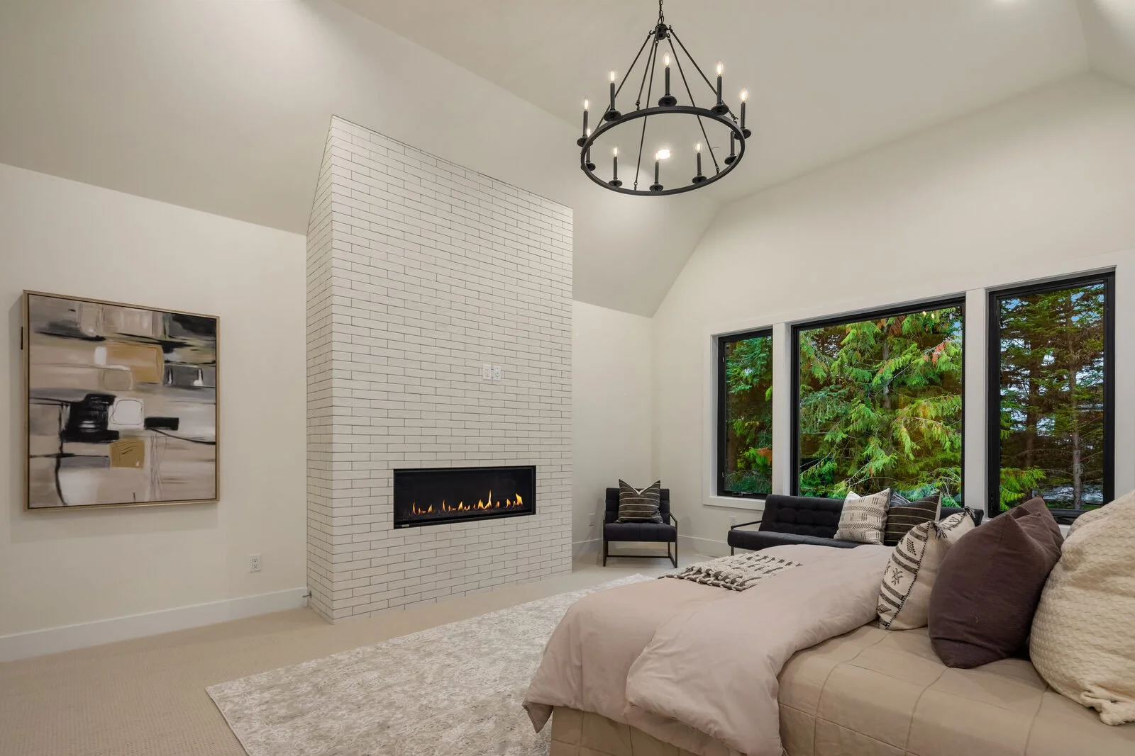 Living room with a white brick fireplace, beige sofa with pillows, black chair, and large windows showing green trees outside. Modern chandelier hangs from ceiling.