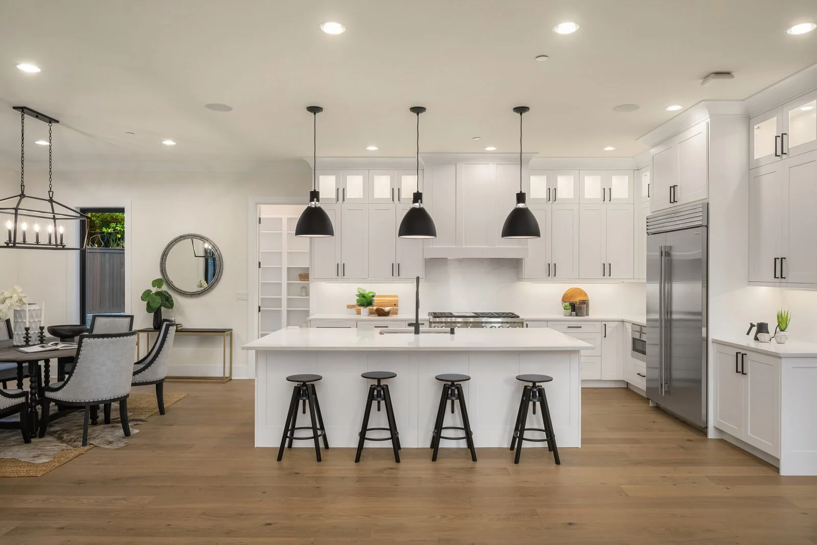 Modern kitchen with white cabinets, black pendant lights over a white island, and a stainless steel refrigerator. Adjacent dining area with table and chairs, decor includes a mirror, potted plants, and a chandelier.