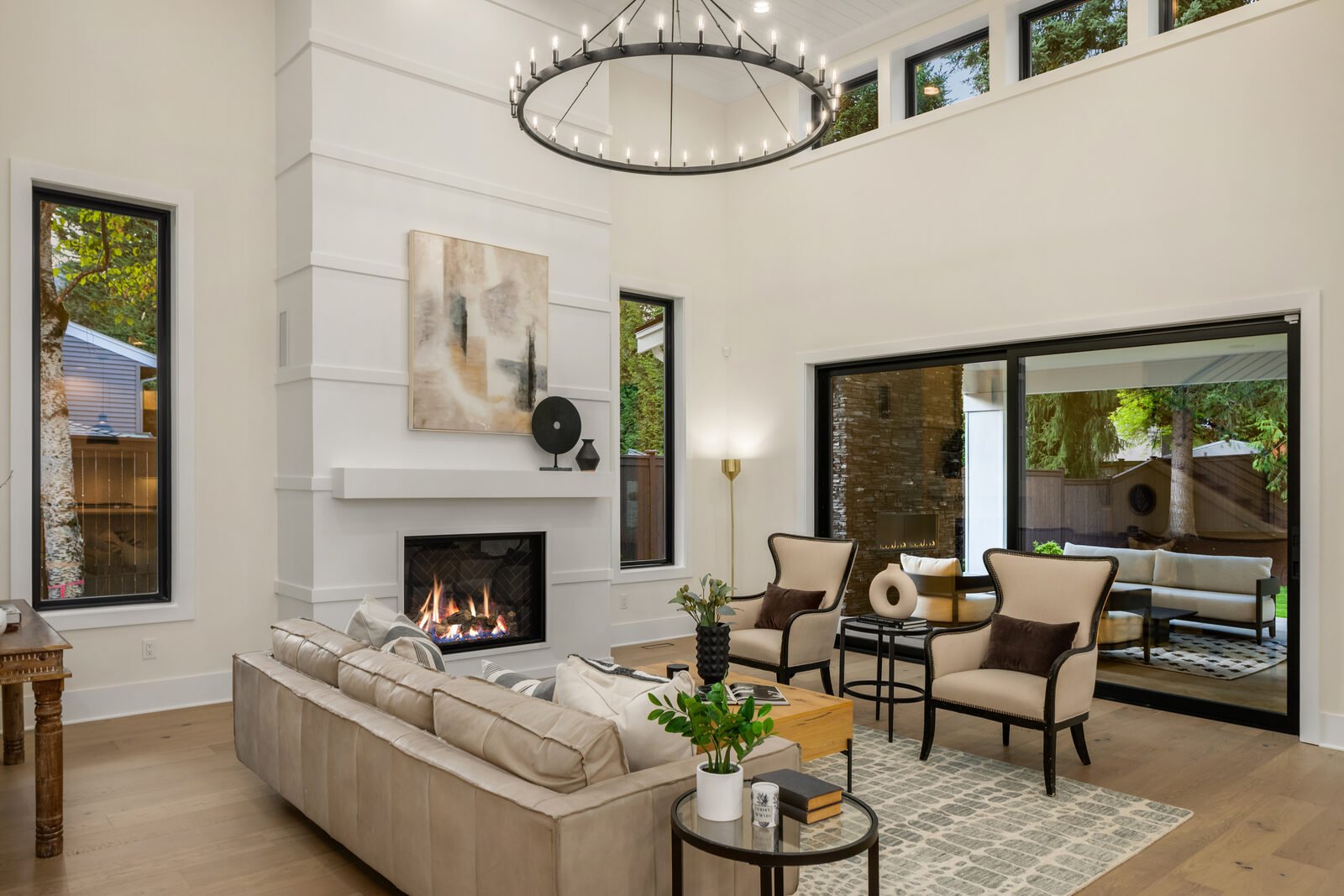Modern living room with white walls, large windows, a fireplace, beige sofa, and armchairs, with a circular chandelier hanging from high ceiling.