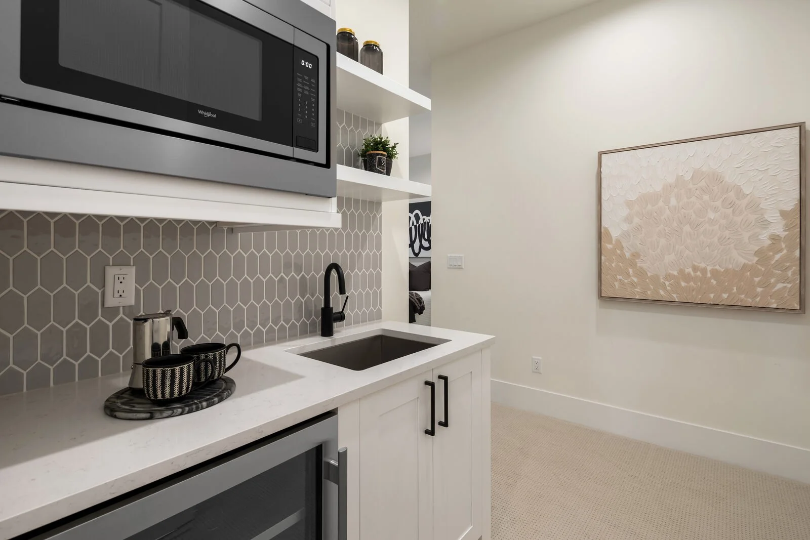 Modern kitchen with white cabinets, black hardware, a black sink with a black faucet, a marble tray with black and white mugs, a speckled granite countertop, gray honeycomb tile backsplash, a microwave oven, a small potted plant, and a large textured
