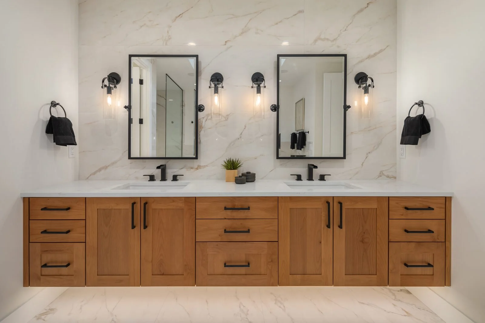 Double vanity bathroom with two mirrors, black fixtures, and black wall-mounted lights. Light wooden cabinets with black handles. Small potted plant and decorative objects on the countertop. White marbled backsplash and walls. Black towels hanging on