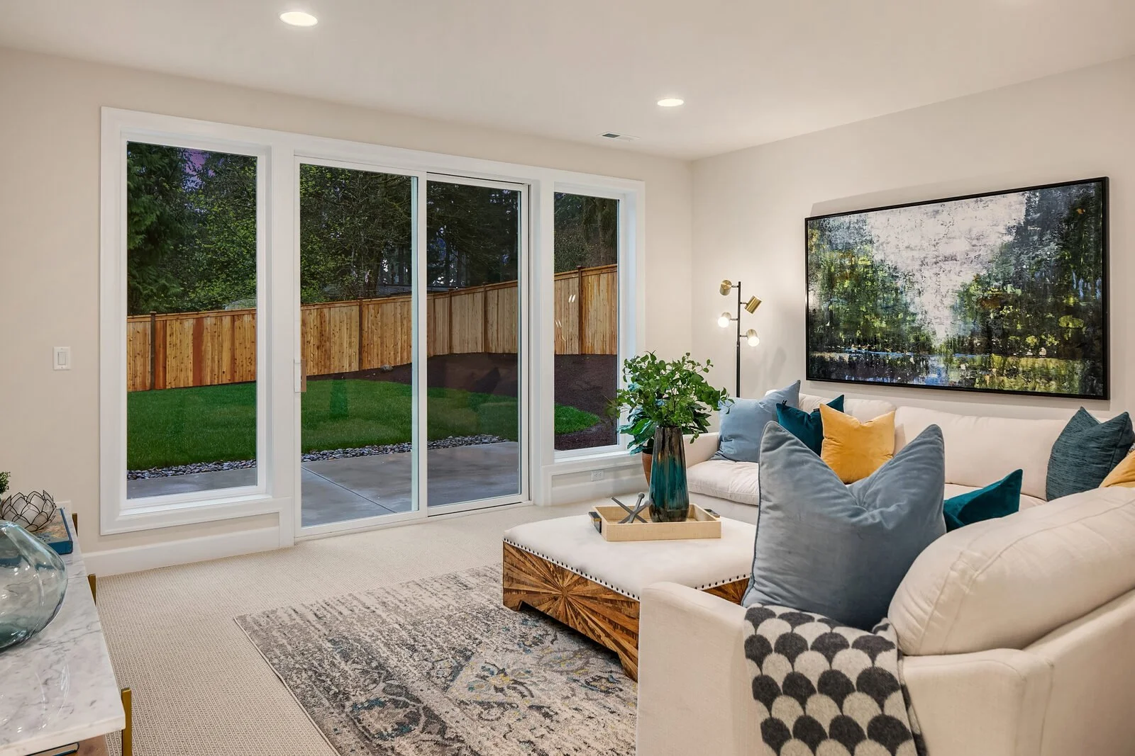 Living room with sliding glass door opening to backyard, white couch with colorful pillows, large wall art, floor lamp, and decorative plants.