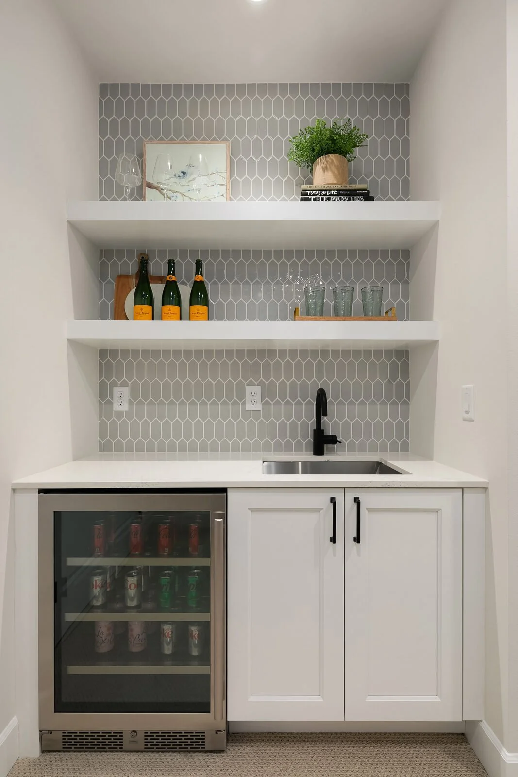 Miniature kitchenette with white cabinets, a black faucet, gray tile backsplash, open shelves with decor and drink bottles, a small bar fridge with beverages, and outlets.