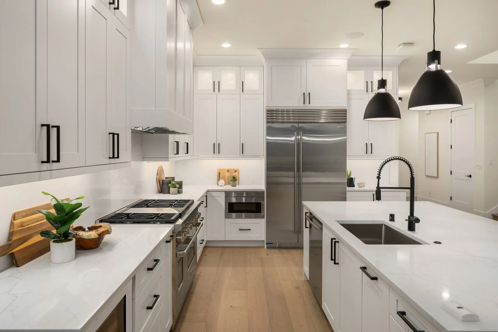 Modern white kitchen with stainless steel appliances, black fixtures, and wooden flooring, featuring a kitchen island with a sink and pendant lighting.