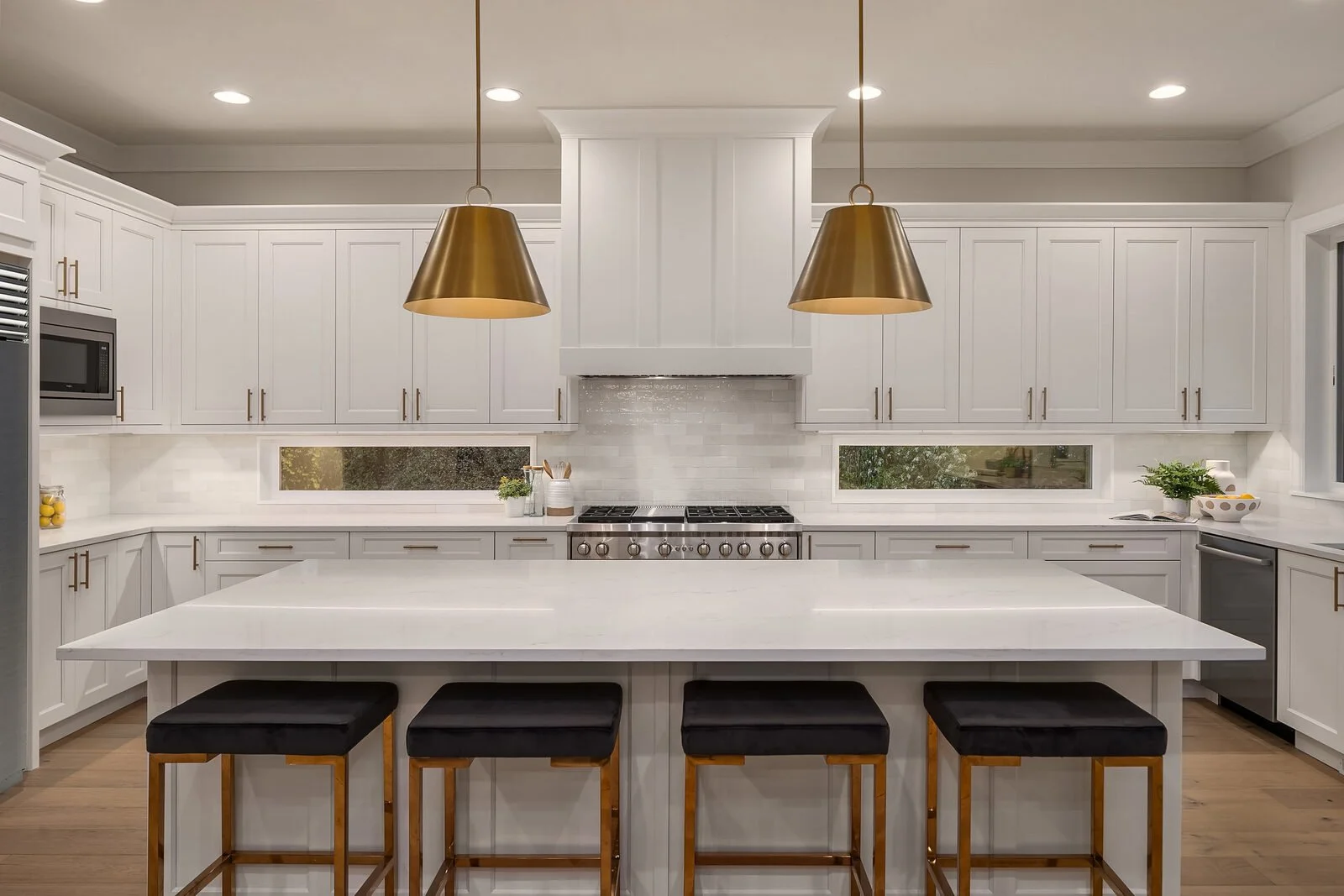 Modern white kitchen with gold pendant lights over a large island, stainless steel appliances, and decorative plants on the countertops.