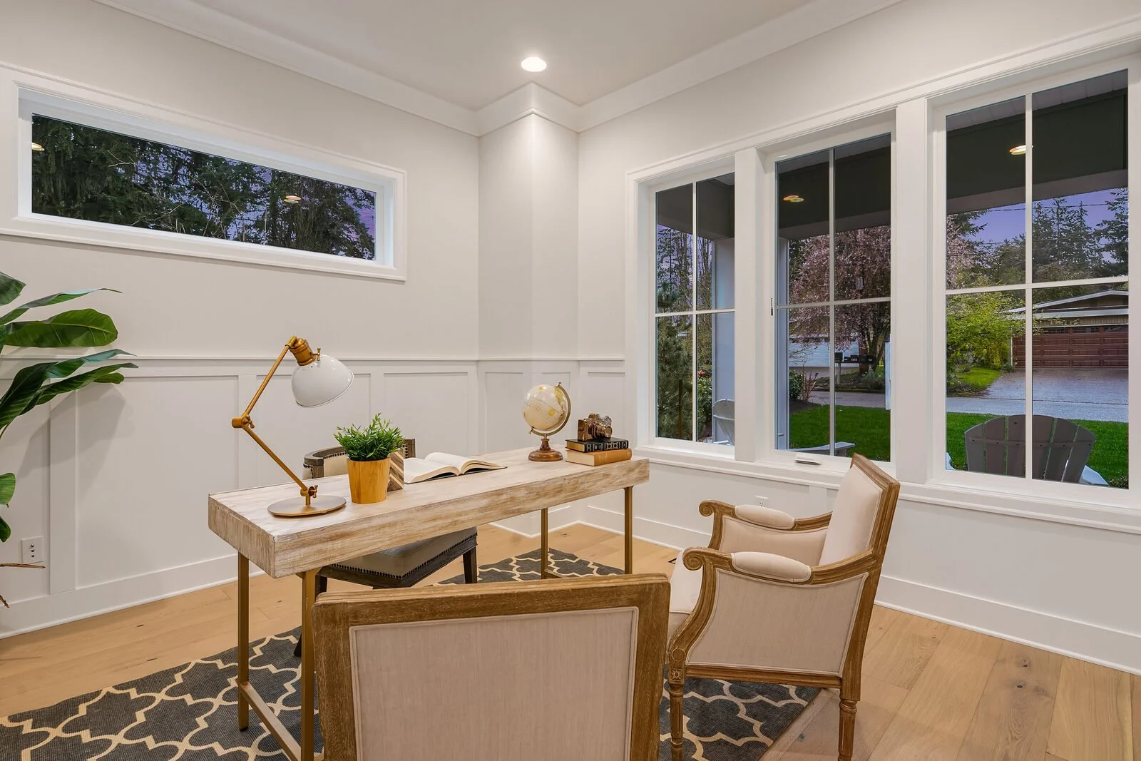 Home office with large windows showing a backyard with trees and a driveway, a wooden desk with a lamp, globe, books, and plant, and two beige chairs.