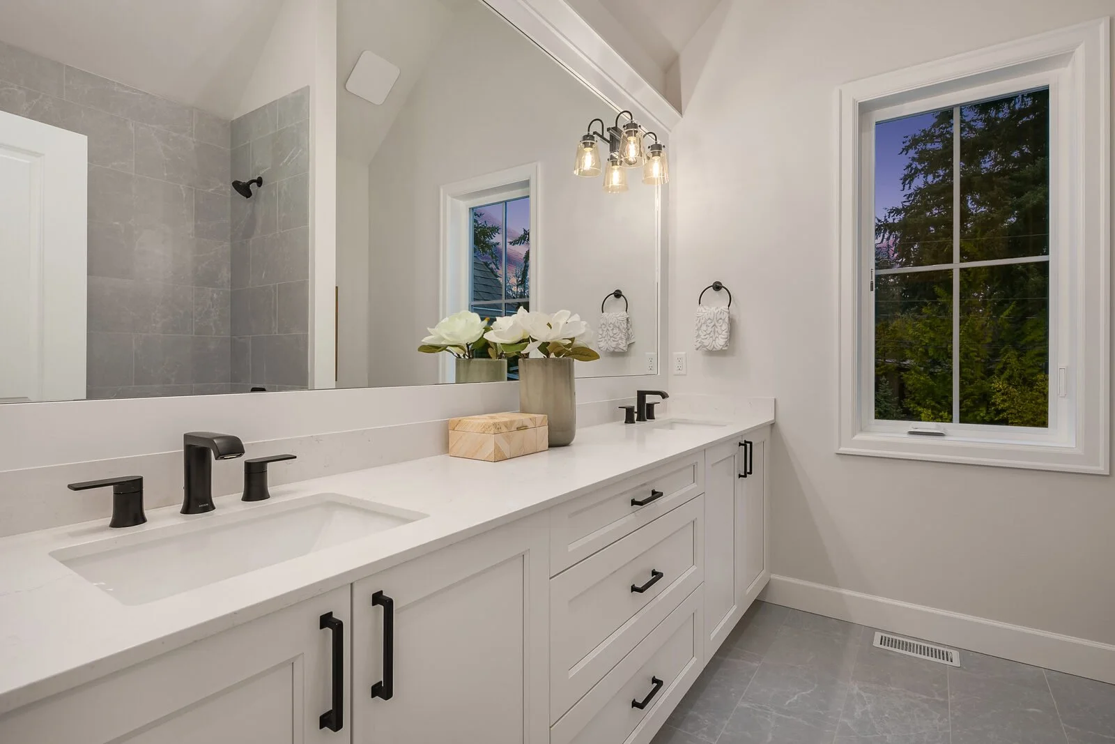 Modern bathroom with a double vanity, black fixtures, a large mirror, and a window showing trees outside. Decor includes a vase with white flowers, towels, and a wooden box.