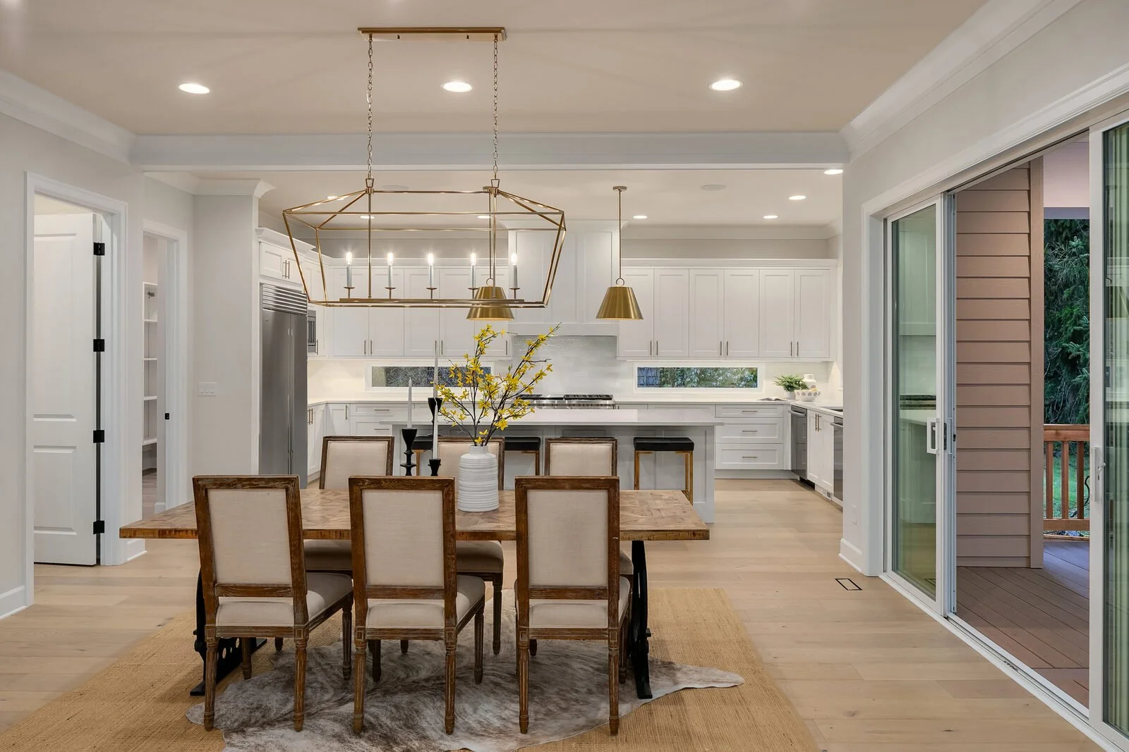 Open-concept dining area leading to white modern kitchen with island and gold pendant lighting, sliding glass door opening to balcony with outdoor greenery.