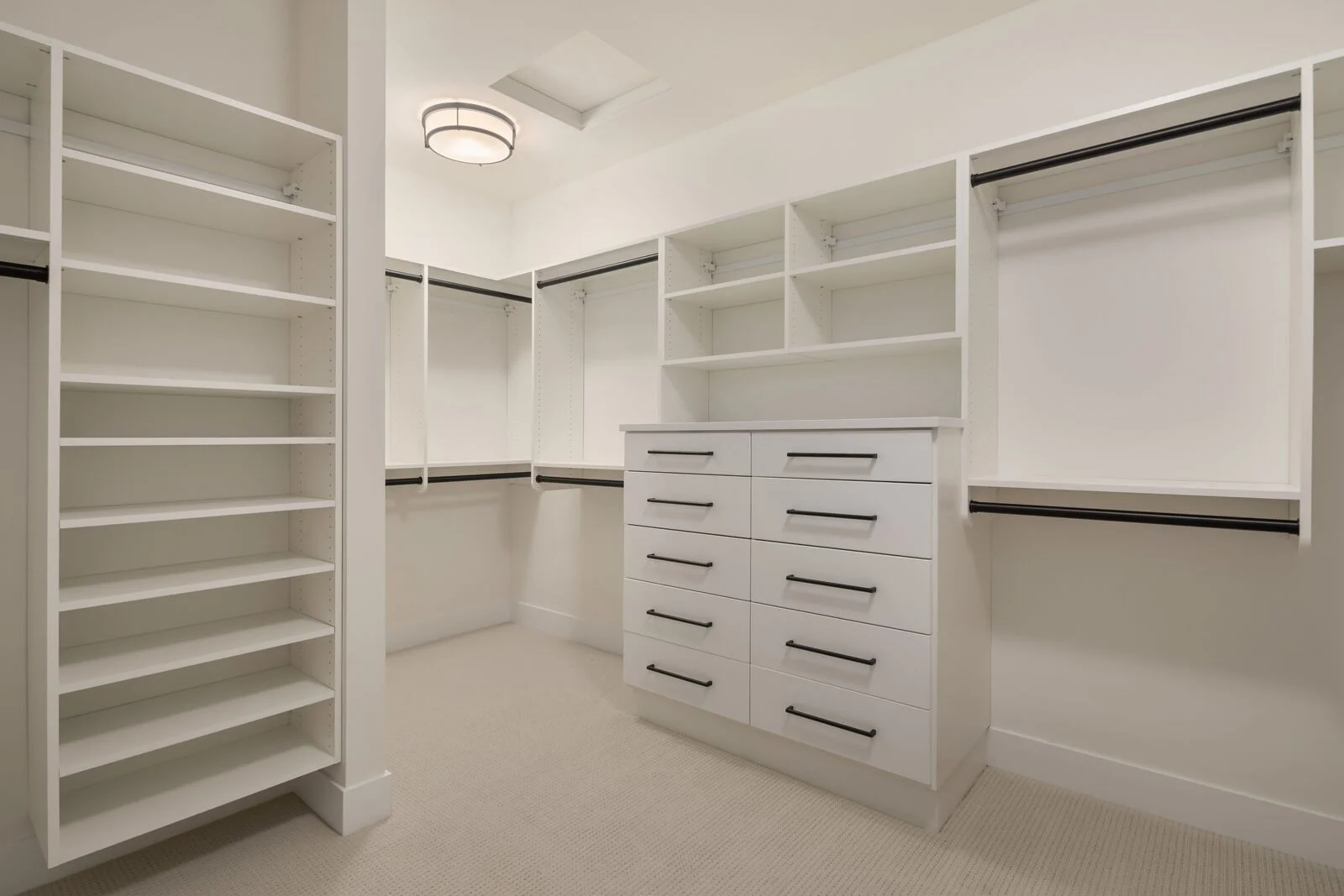 Empty walk-in closet with white shelving and drawers, black hanging rods, and beige carpet flooring.