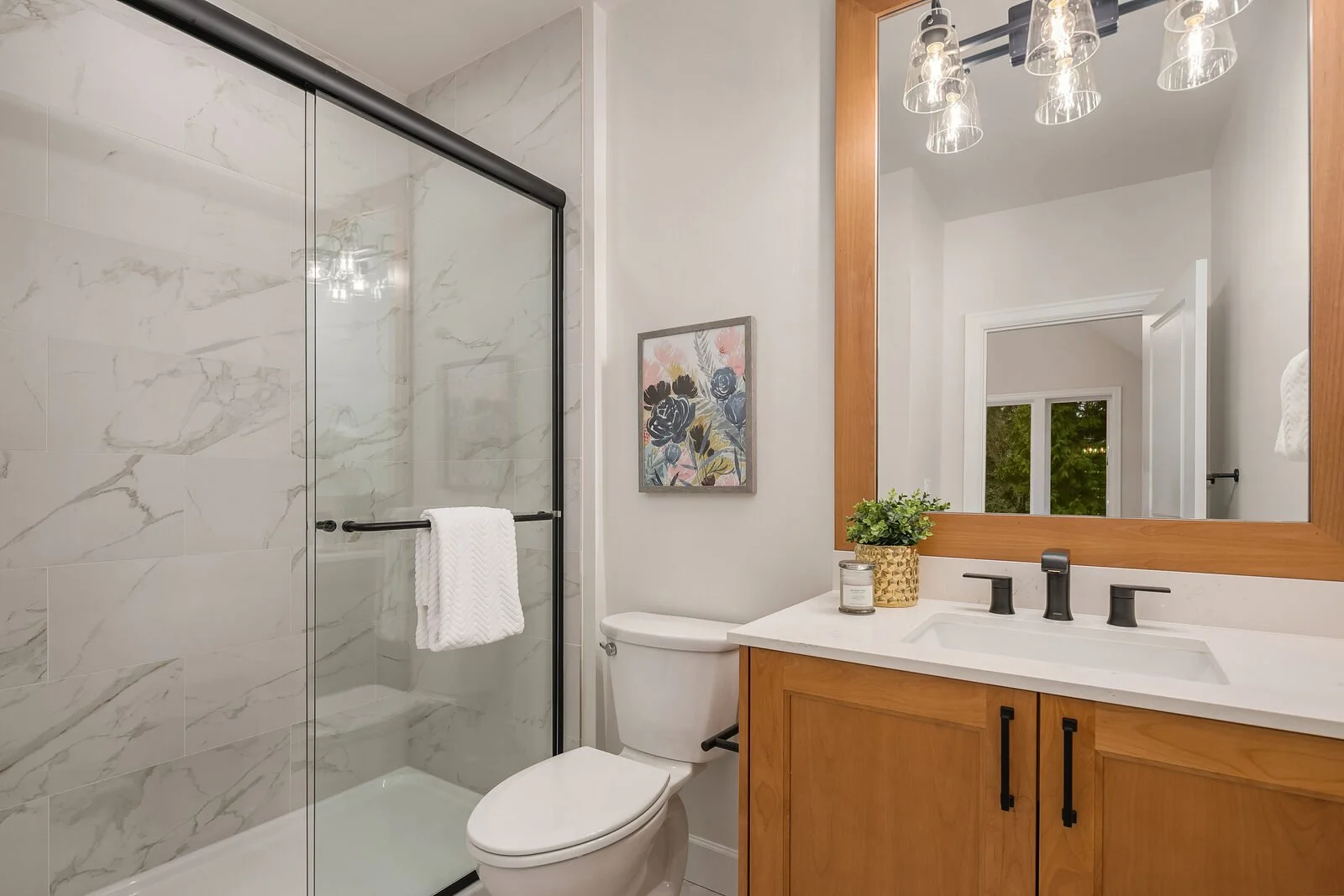 Bathroom with glass shower enclosure, wooden vanity with white countertop, large mirror, and decorative items, including a framed floral picture and small plant.