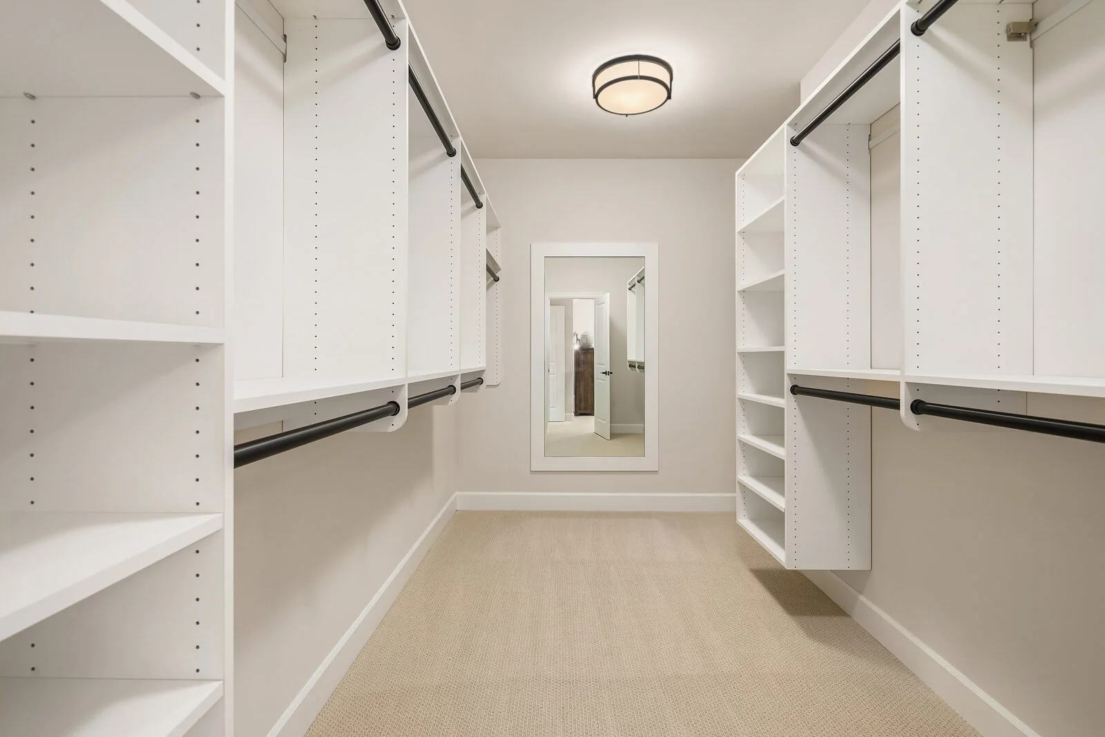 Empty walk-in closet with white shelves and hanging rods, carpeted floor, and a large mirror on the back wall.