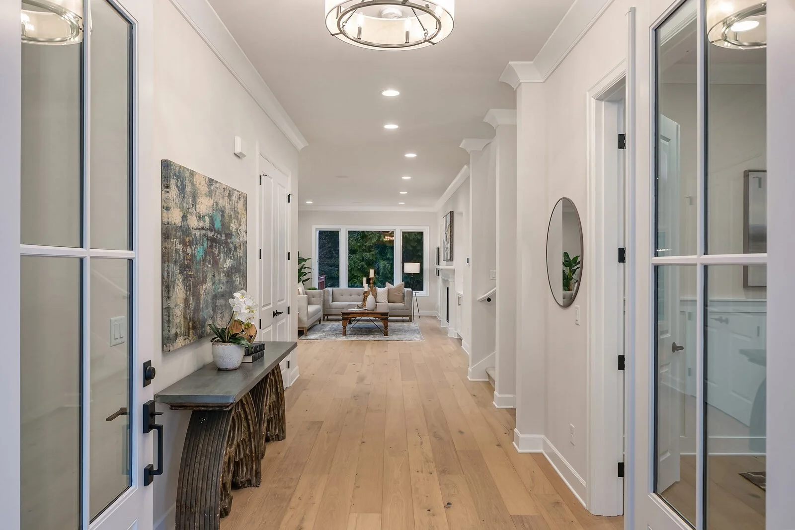 A bright living room with white walls and ceiling, hardwood floors, a modern chandelier, and recessed lighting. The room has a sitting area with white sofas, a wooden coffee table, and a large window showing greenery outside.
