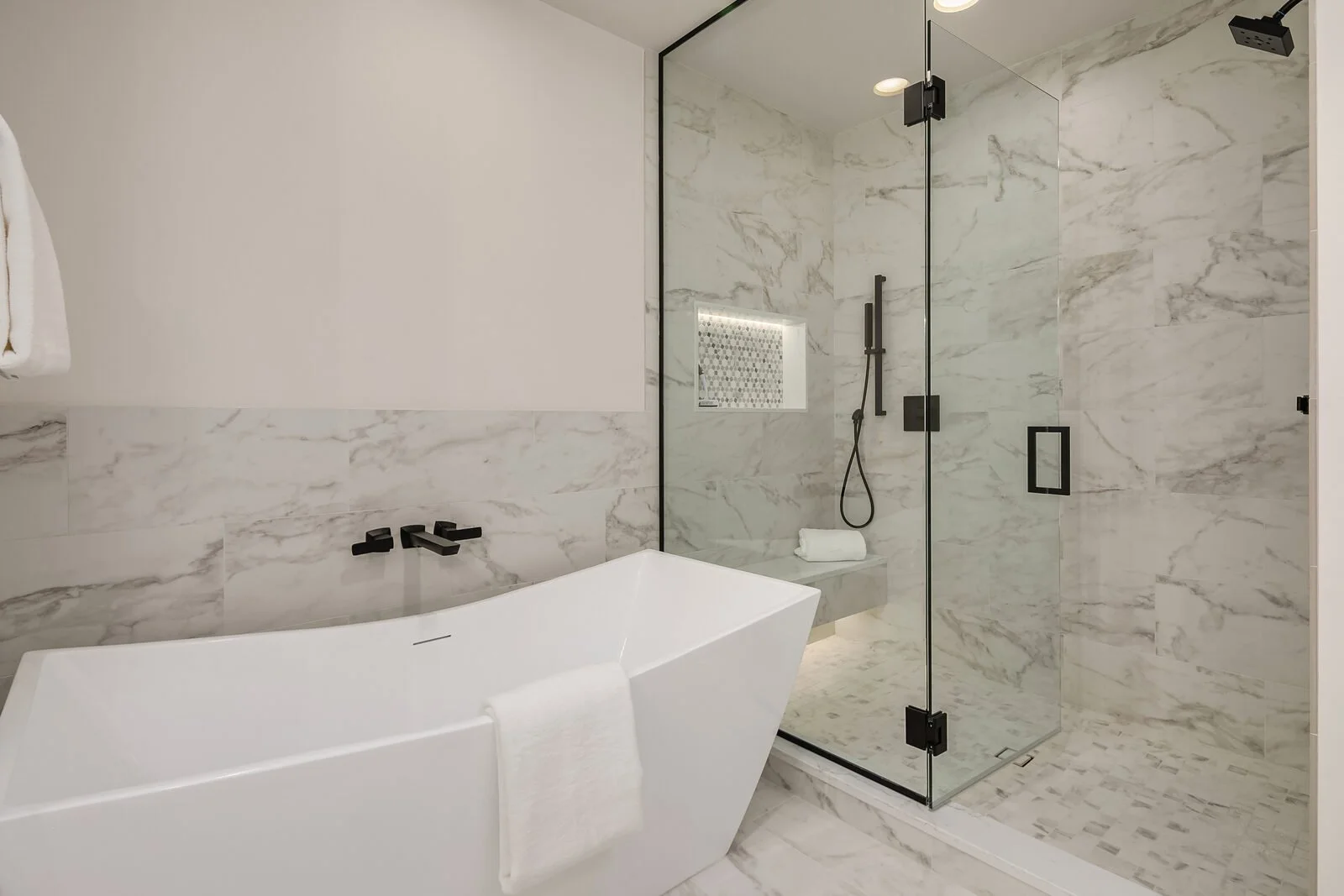Modern bathroom with white freestanding tub, black fixtures, and a separate walk-in glass shower with marble walls and floor.