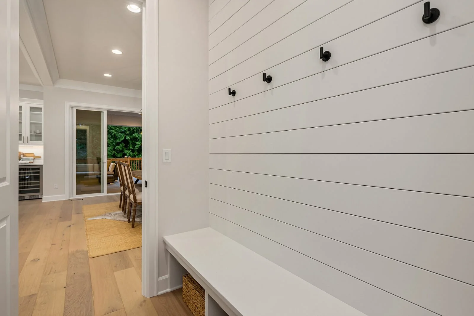 White shiplap wall with black hooks and a white bench underneath. A doorway leads to a dining area with a table, chairs, and a sliding glass door to an outdoor deck with greenery.