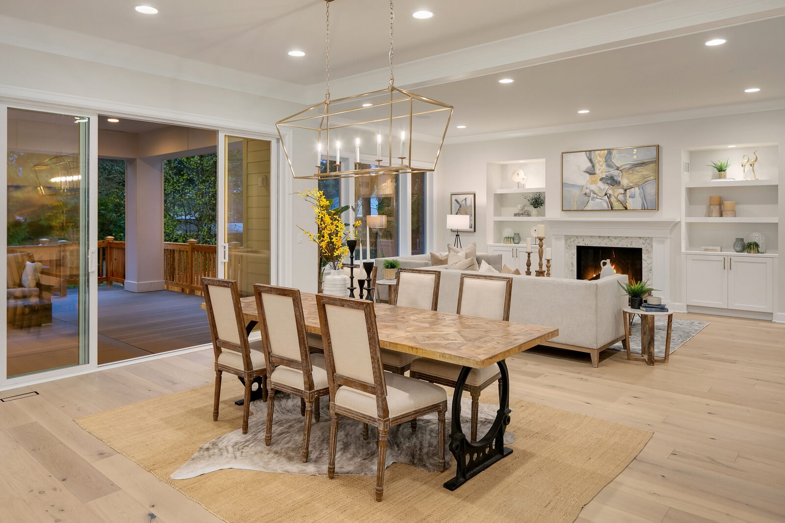 Open-concept living and dining room with a wooden dining table, cream upholstered chairs, white sectional sofa, fireplace, mounted artwork, built-in shelves, and a sliding glass door leading to a balcony with outdoor seating.